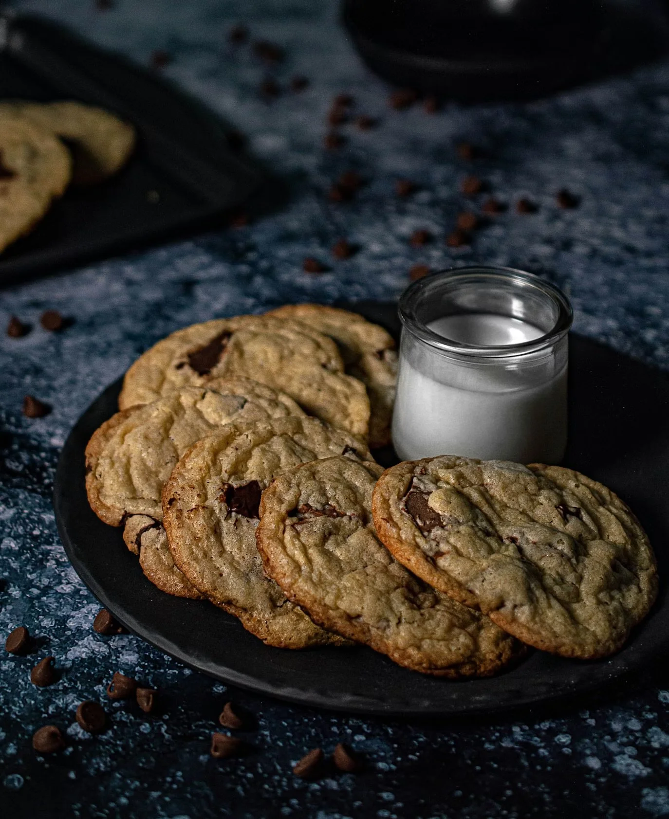 Chocolate chip cookies on a black plate with a small glass of milk on dark textured surface.