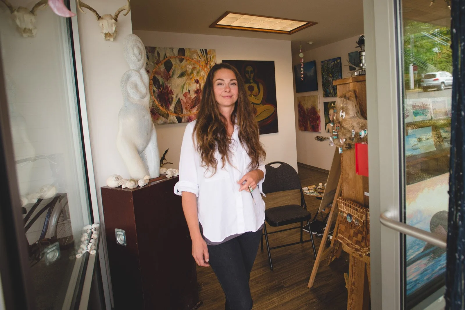 Andrea Fergusson standing in art gallery, surrounded by paintings and sculptures. She is wearing a white shirt and dark pants, with long wavy brown hair, smiling at the camera.
