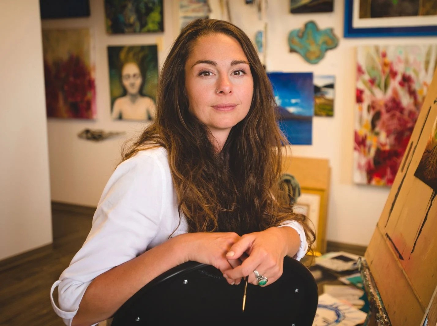 A woman with long, wavy brown hair and a nose ring, wearing a white shirt, sitting at an easel in an art studio surrounded by various colorful paintings on the wall.