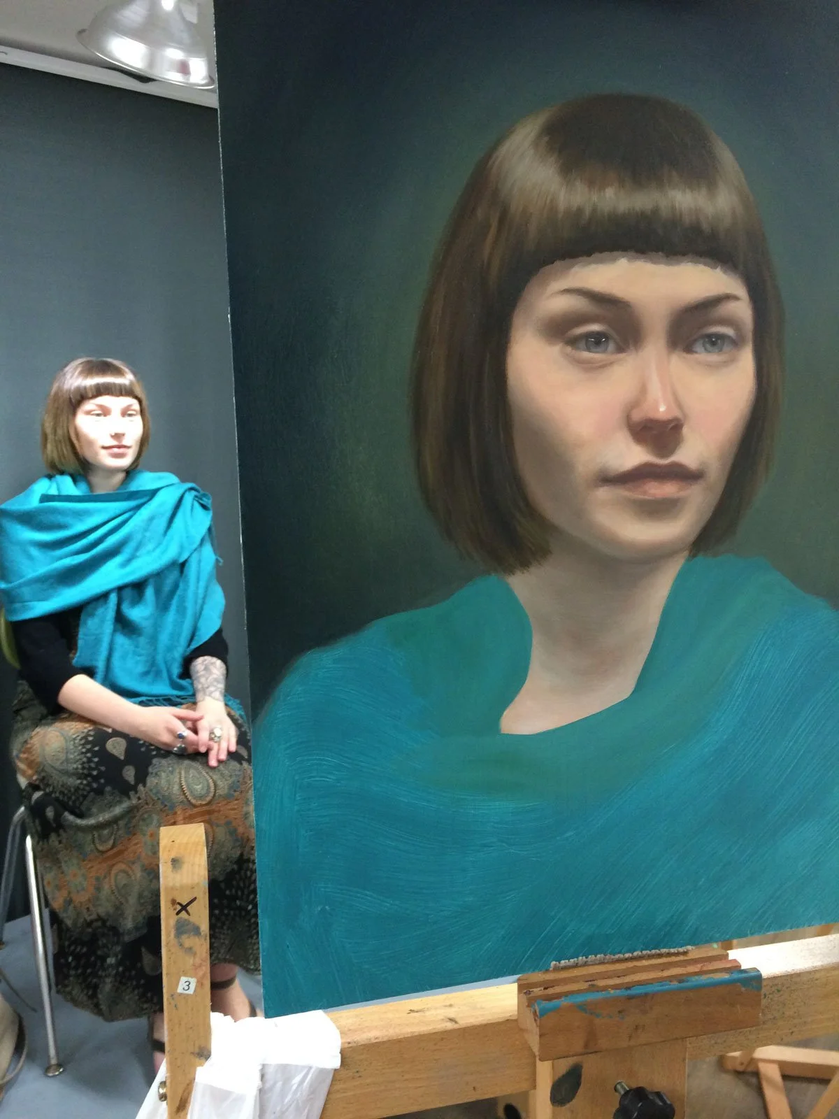 An artist's painting of a woman with short brown hair and a teal top, displayed on an easel, with a woman sitting in the background.