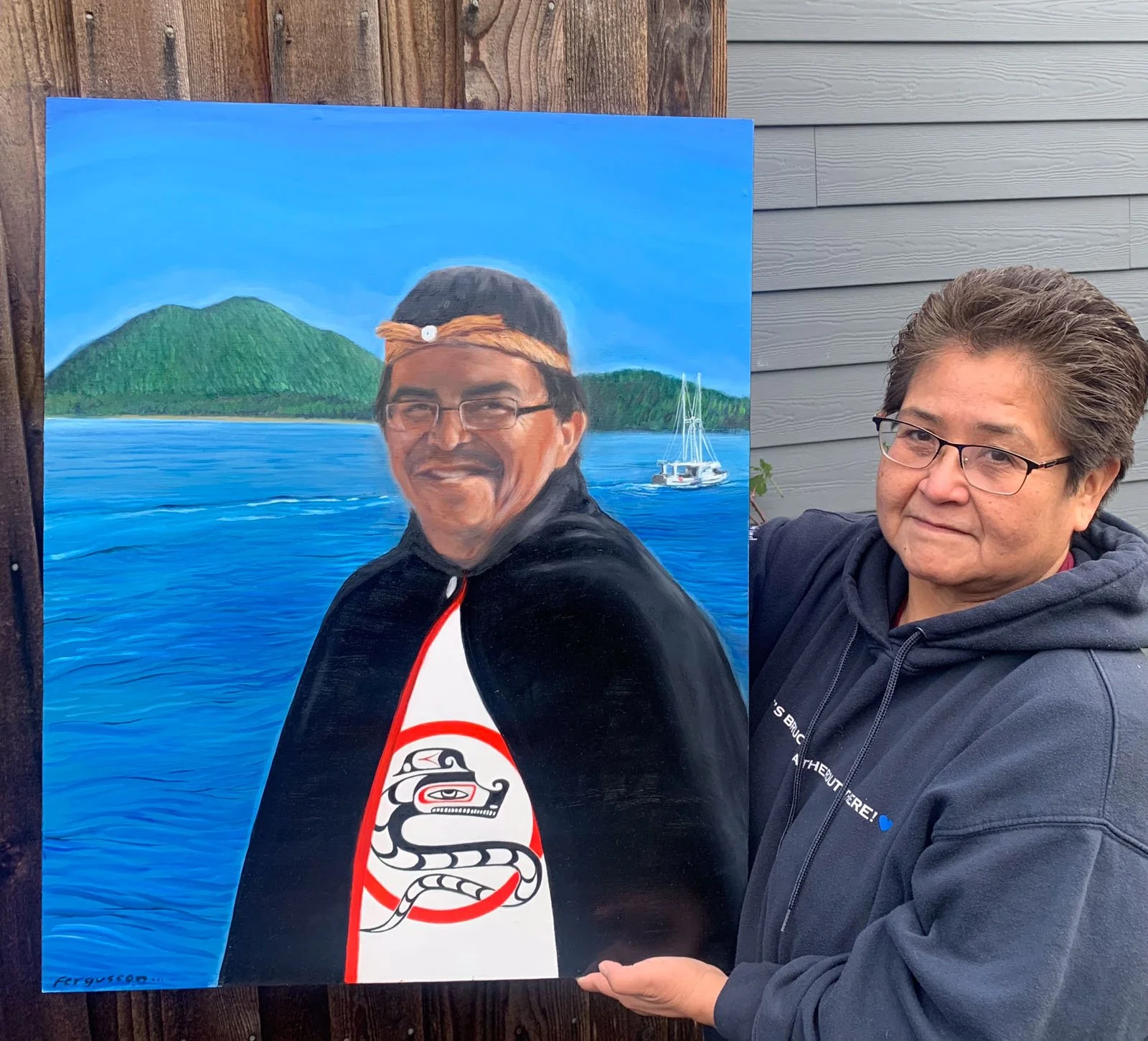 A woman holding a colorful painting of a man with glasses, wearing a black jacket and a headband, standing outdoors near a wooden fence. The painting depicts a lakeside scene with a sailboat and green hills in the background.