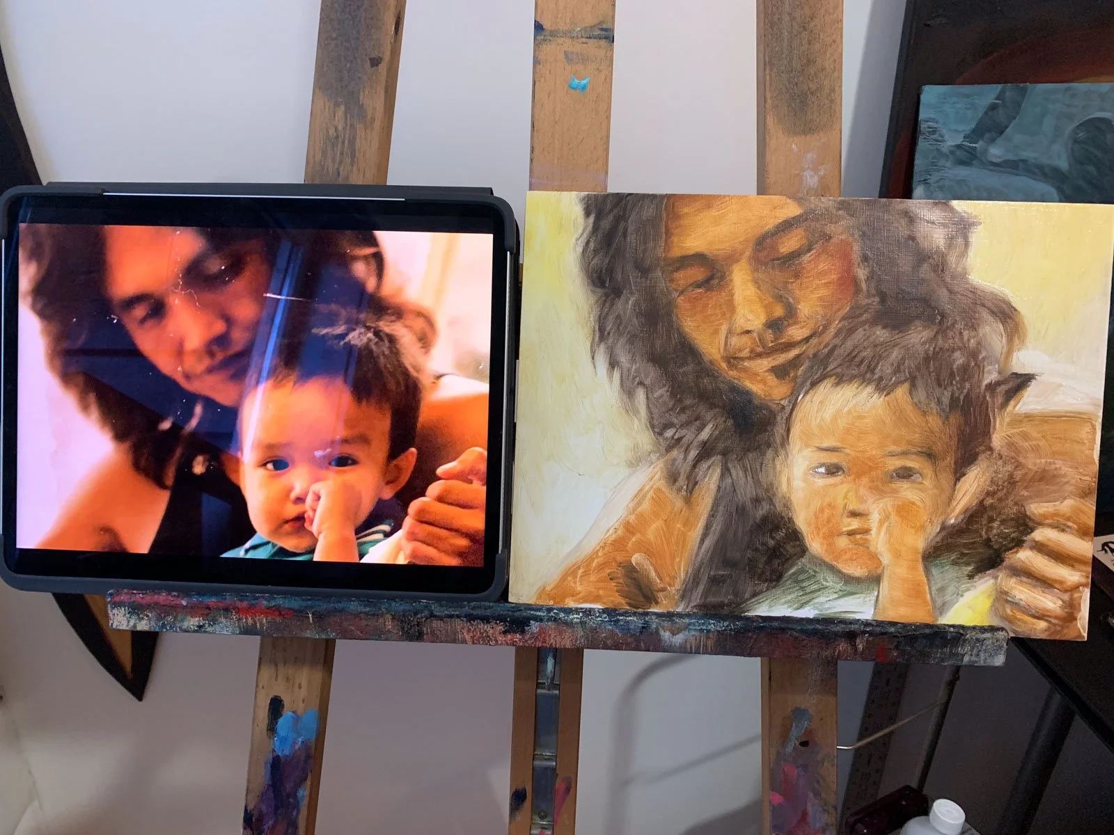 A digital tablet with a photo of a woman and a young boy pausing from a painting of the same woman and boy that is on an easel.