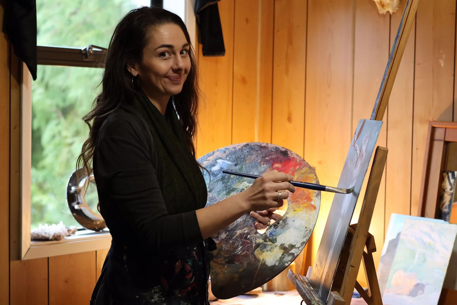 Woman with dark hair smiling while painting on a canvas in an art studio with wooden walls