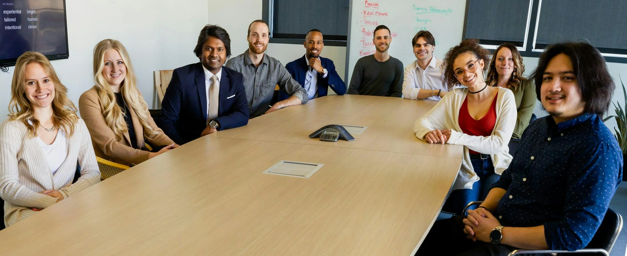 A diverse group of ten people sitting around a conference table in a modern office, smiling at the camera.