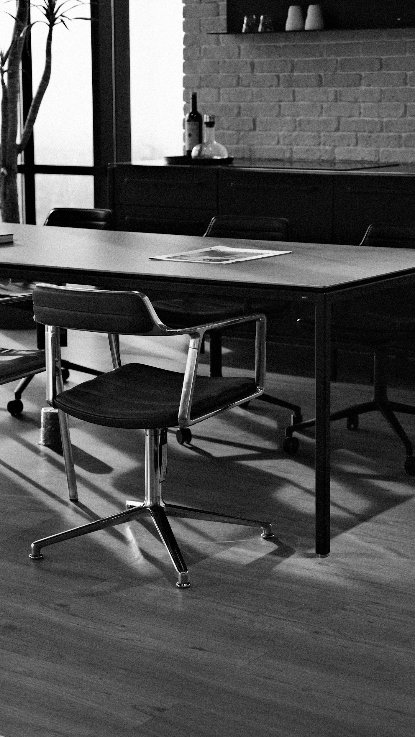 A black and white photo of a modern meeting room with a wooden table, rolling office chairs, and a brick wall with a shelf holding decorative items.