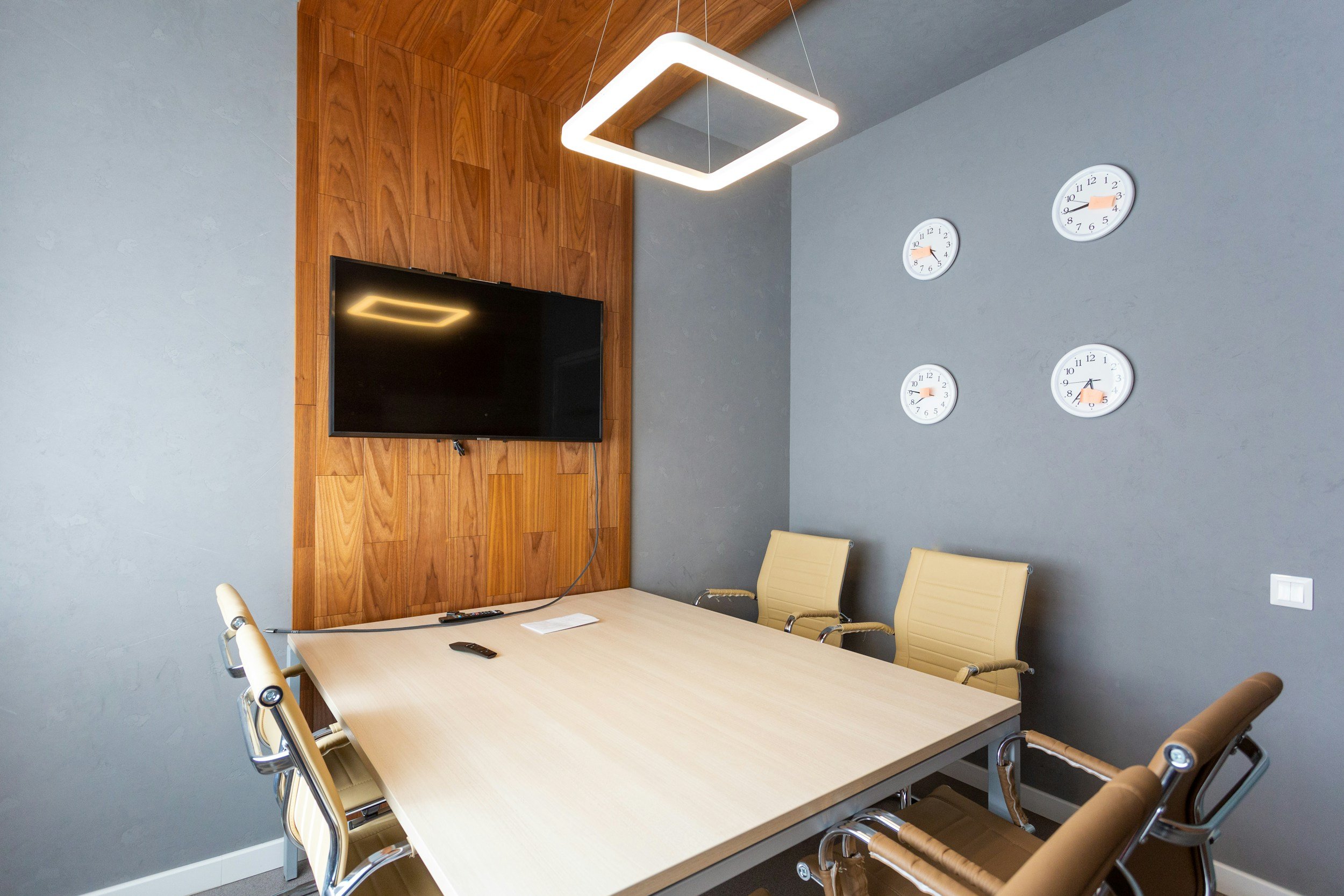 Modern conference room with a light wood table, six beige office chairs, a wall-mounted TV, four clocks on the wall, and a square LED ceiling light.