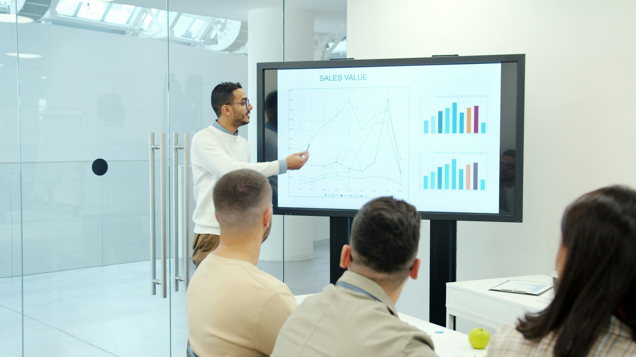 A man is giving a presentation to a group of people in a modern, glass-walled conference room with a big screen showing sales charts.