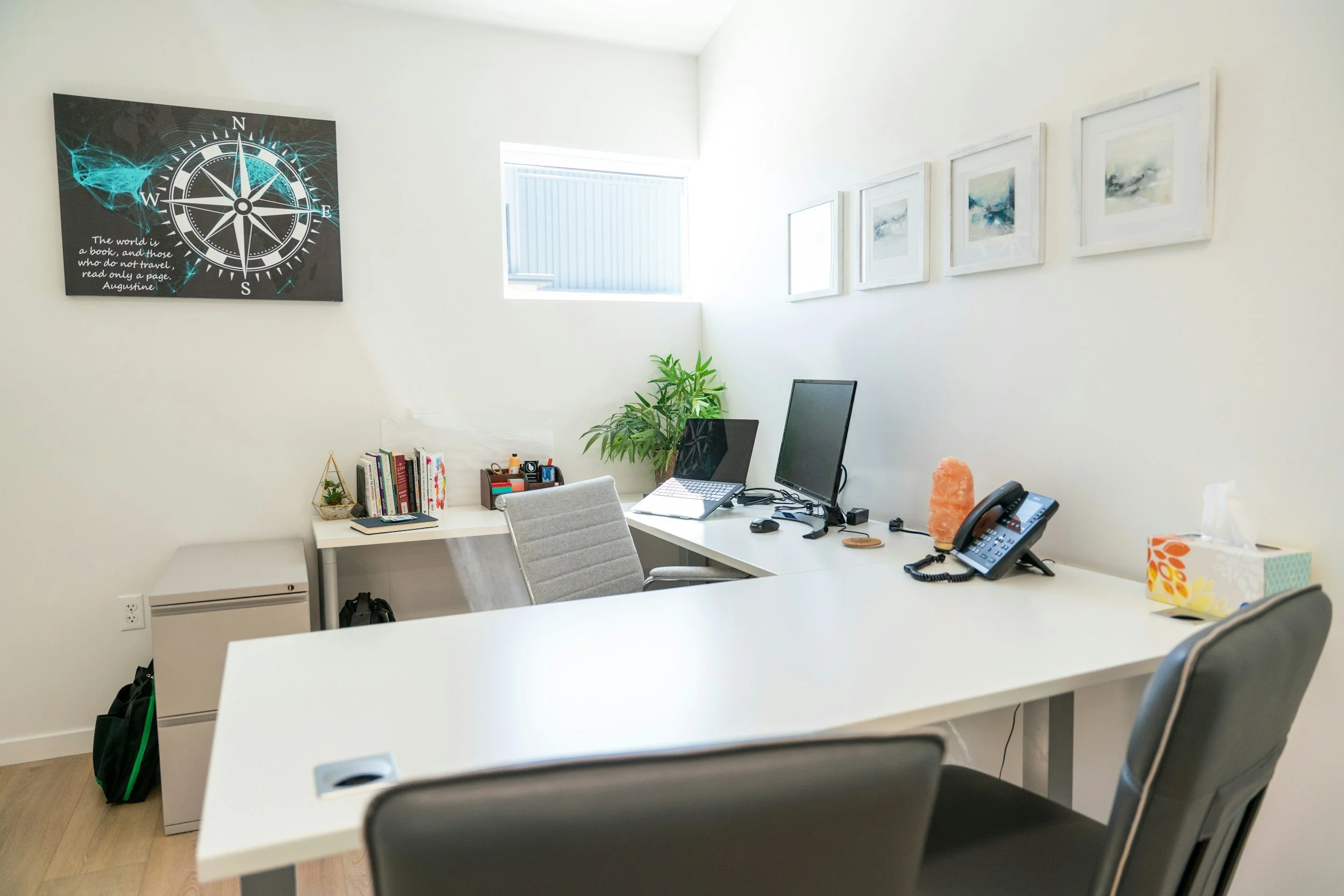 Modern office with white walls, a white desk, black monitor, office phone, framed artwork on the wall, a window with natural light, a small potted plant, and office supplies.