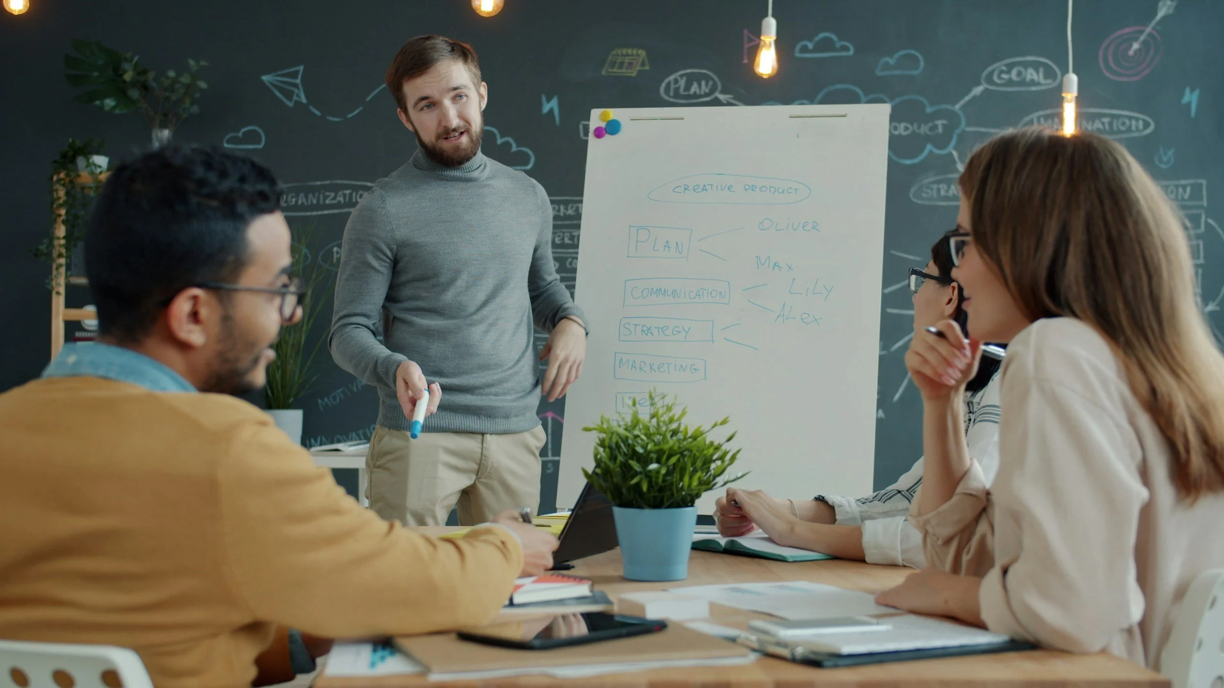 Man giving presentation to group of people sitting at a table in an office setting with a whiteboard and chalkboard in the background.