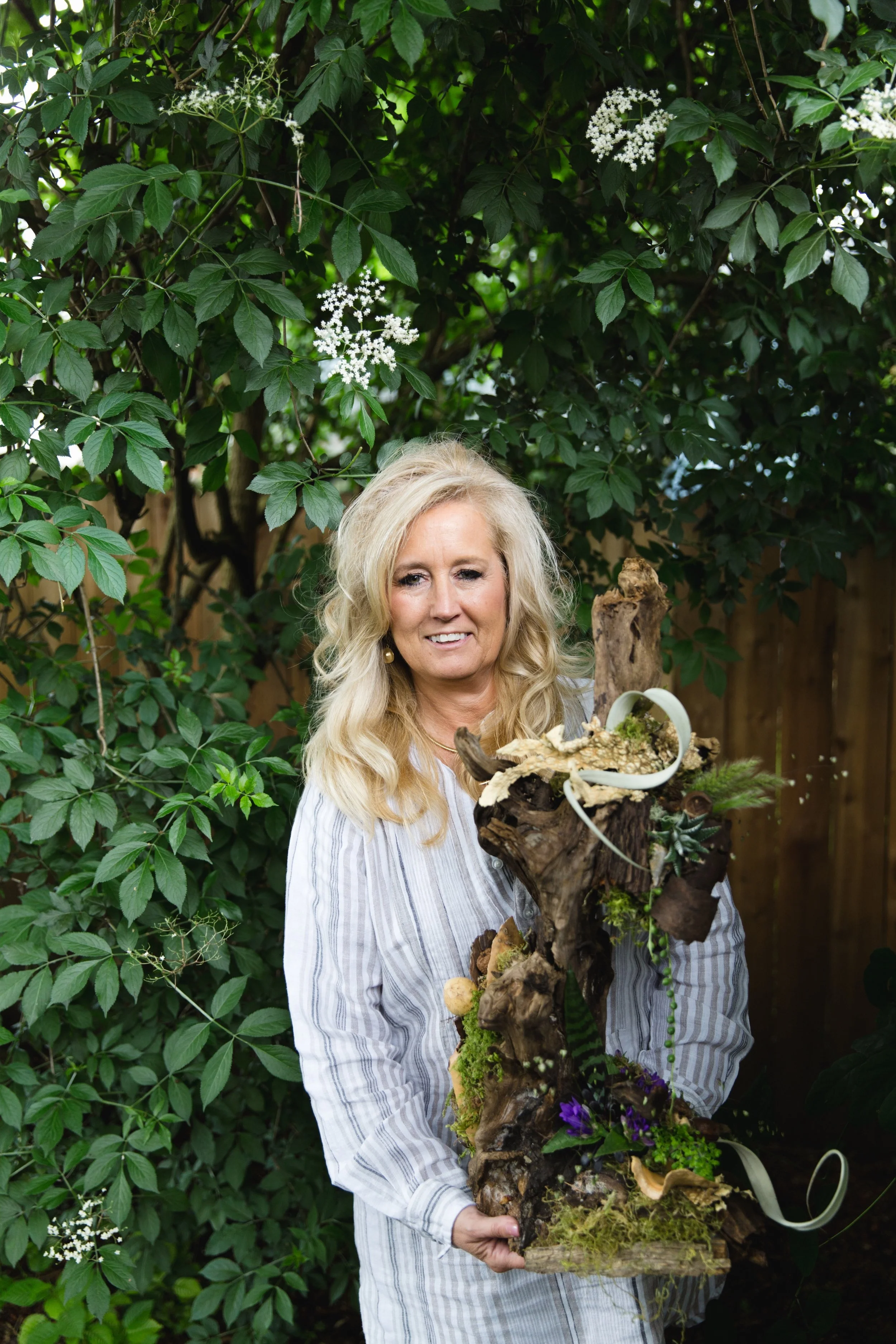 A woman with blonde hair wearing a striped gray and white shirt holding a nature-inspired sculpture outdoors amid green foliage and white blossoming flowers.