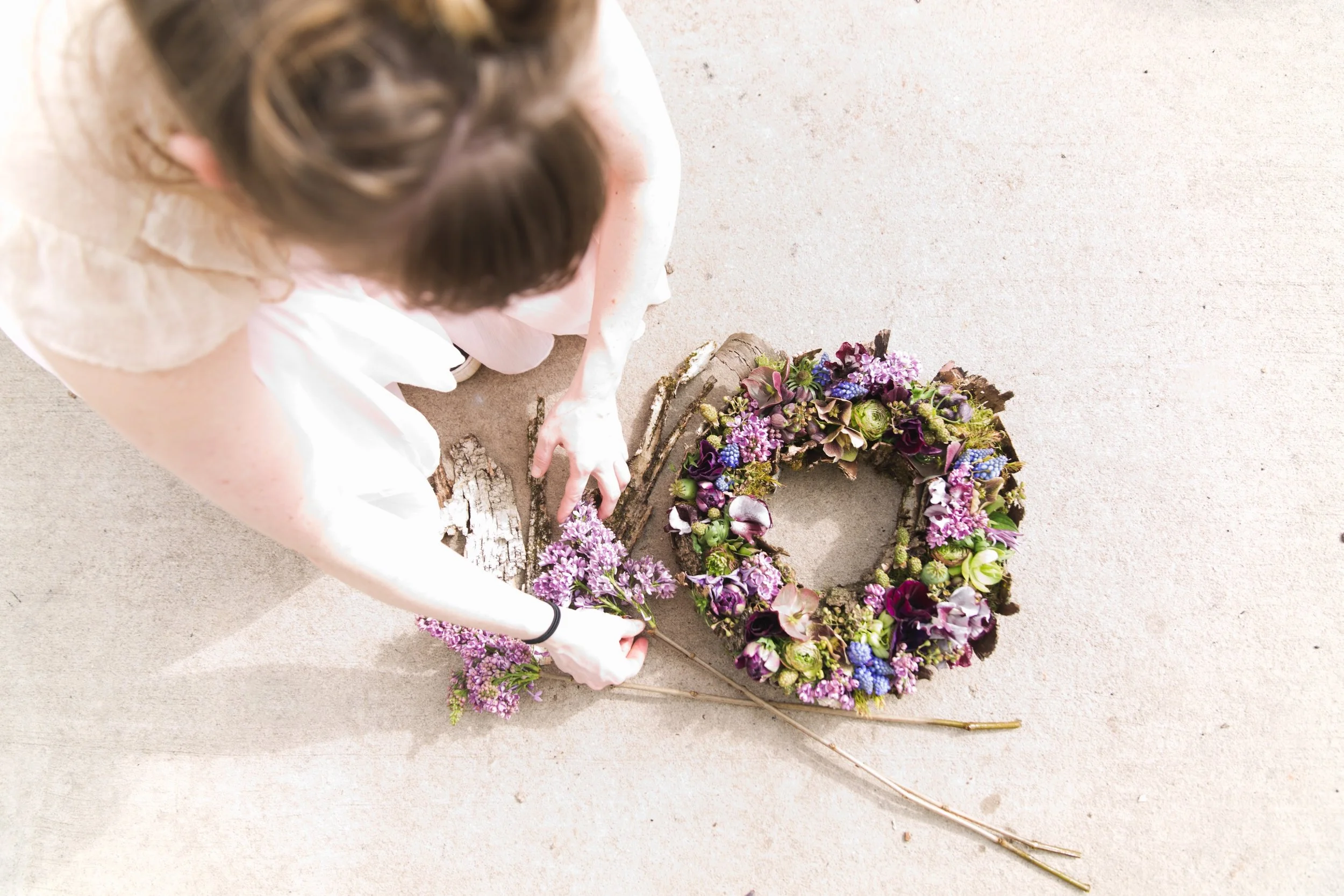 A woman arranging a flower wreath on a beige floor, with two long stems of purple flowers nearby.