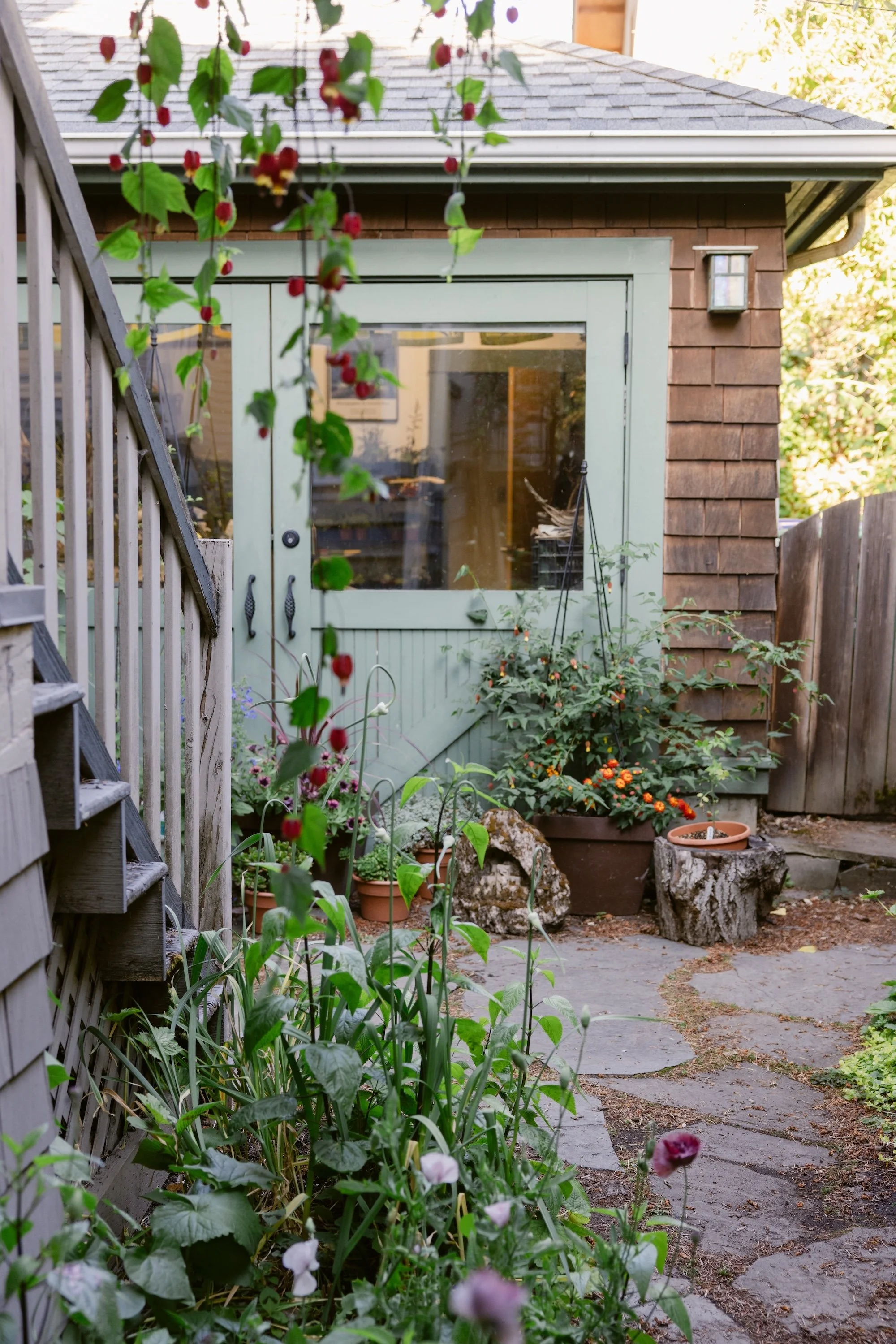 A backyard garden with potted plants and flowers, a stone path, and a studio with a glass door.