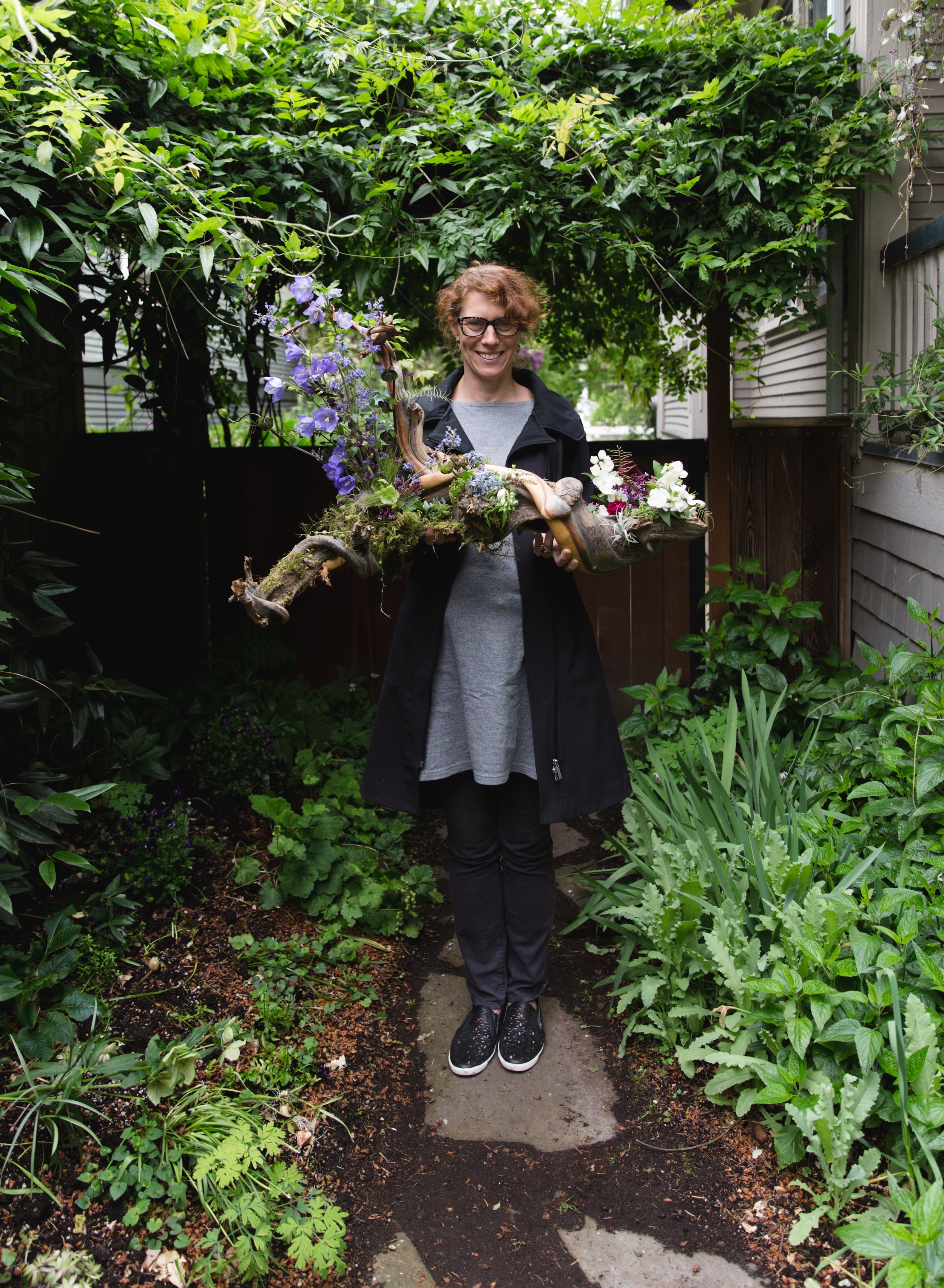 A woman standing on a garden pathway holding a large wooden and floral arrangement, surrounded by green plants and trees.