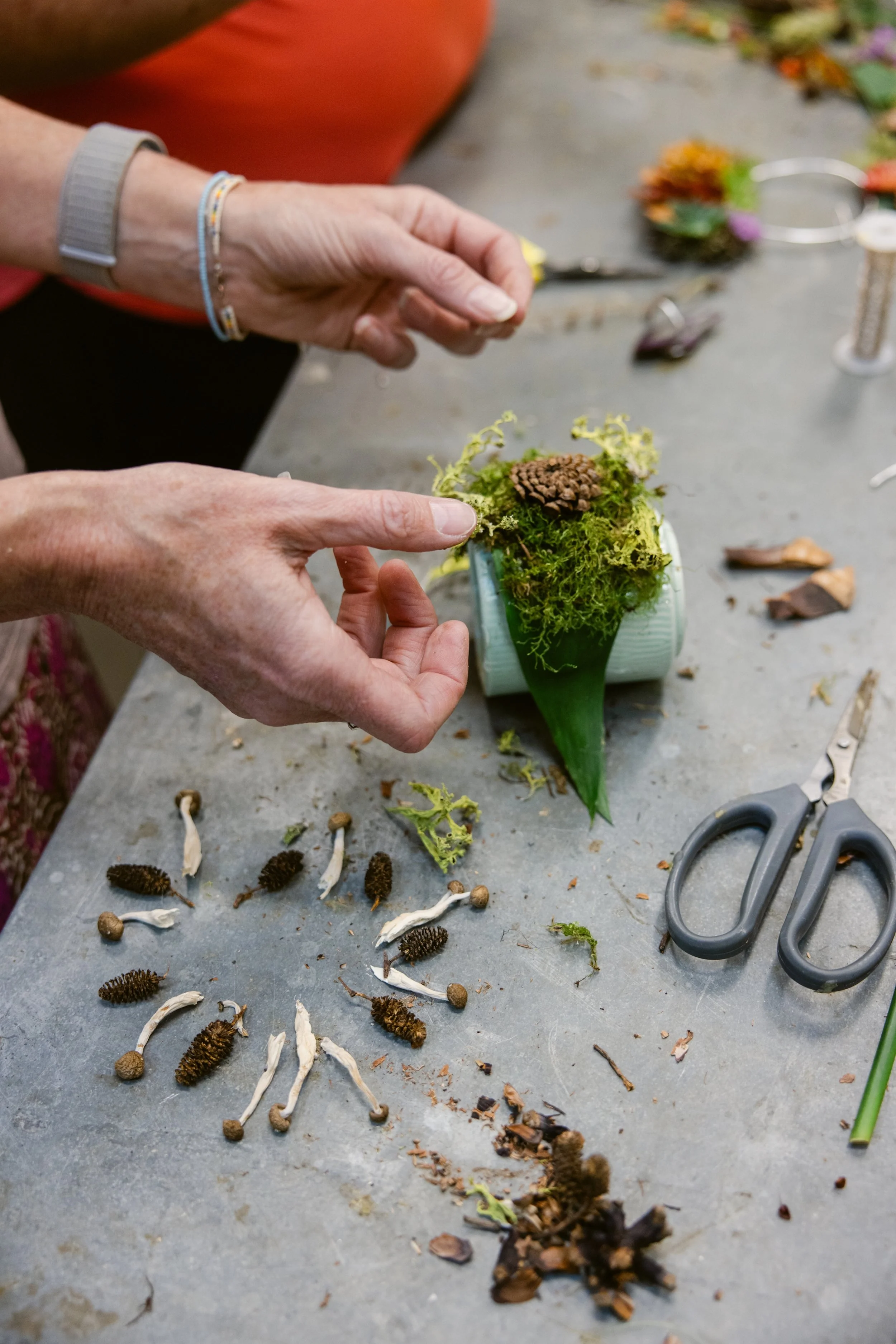 Close-up of hands assembling a botanical arrangement with moss, leaves, and other natural elements on a worktable, with scissors and scattered natural materials around.