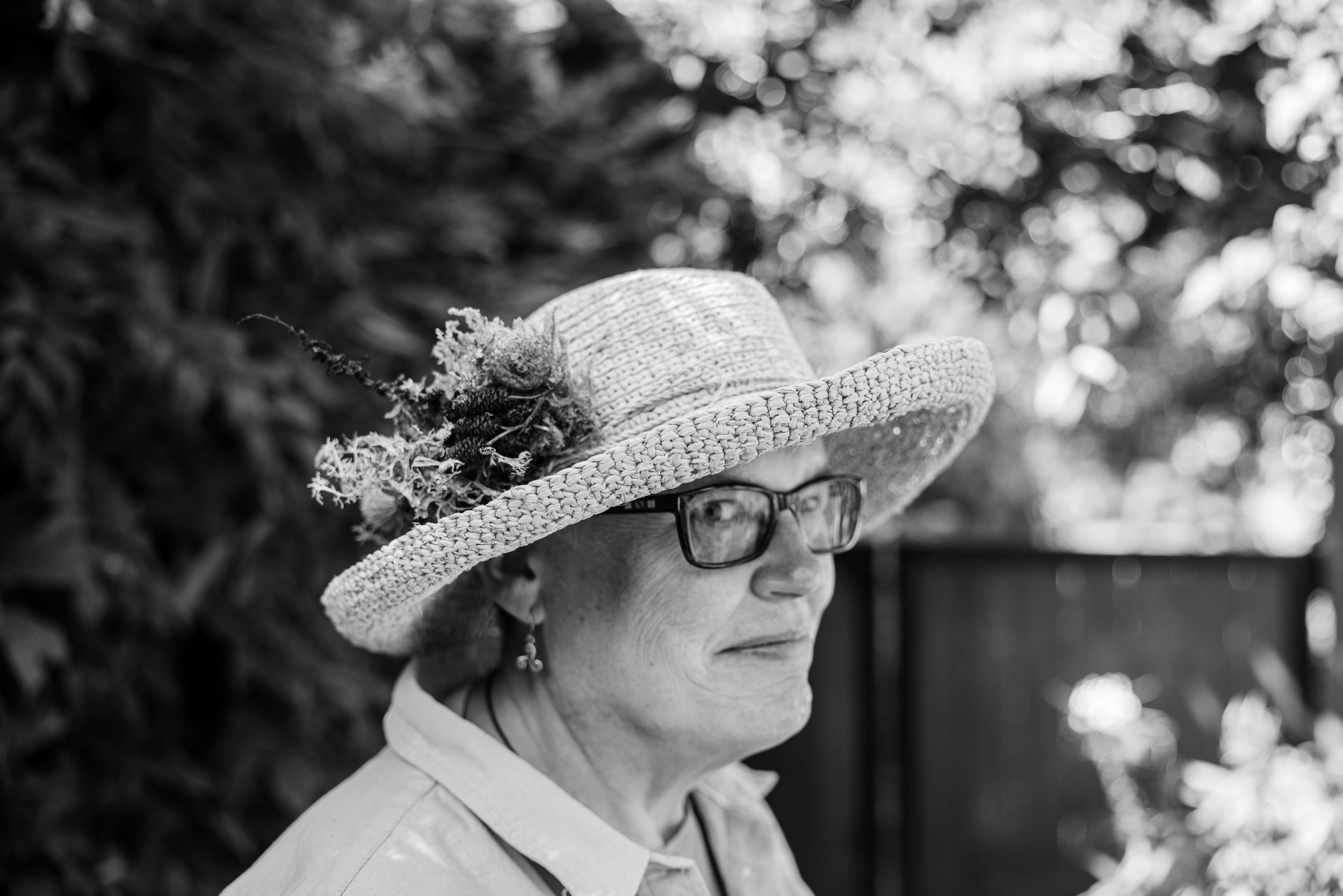 A grayscale photo of an elderly woman wearing a wide-brimmed straw hat decorated with flowers and a large flower pin, glasses, and earrings, standing outdoors with blurred foliage in the background.