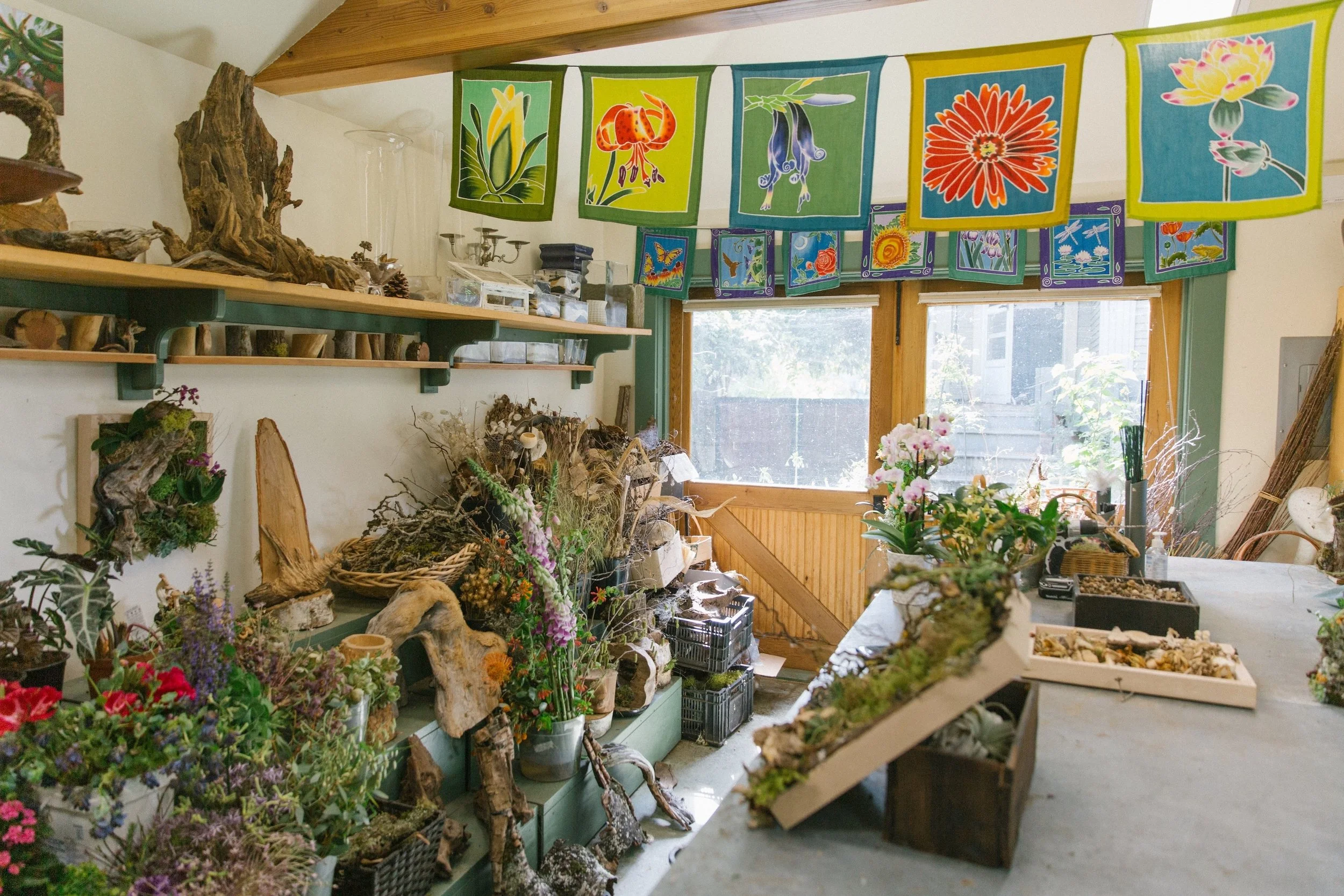 A room decorated with colorful flower-themed banners hanging from the ceiling, shelves displaying natural wood and plant arrangements, and a work table with flower and plant supplies, with a large window letting in sunlight.