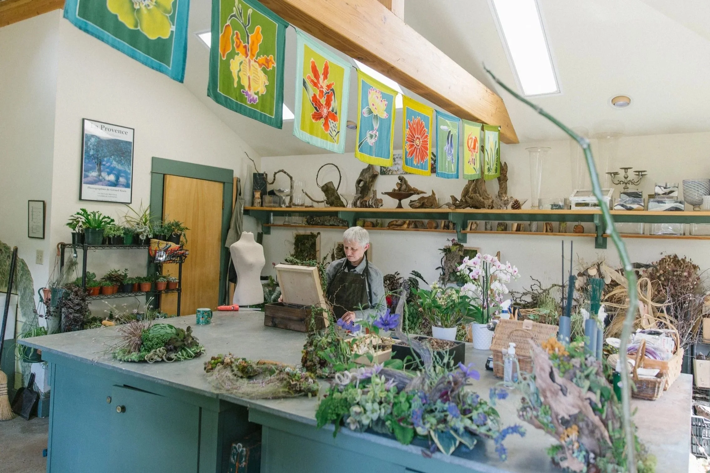 An artist working on floral arrangements or botanical art in a cozy studio filled with plants, flowers, and natural decor, with colorful fabric banners hanging from the ceiling.