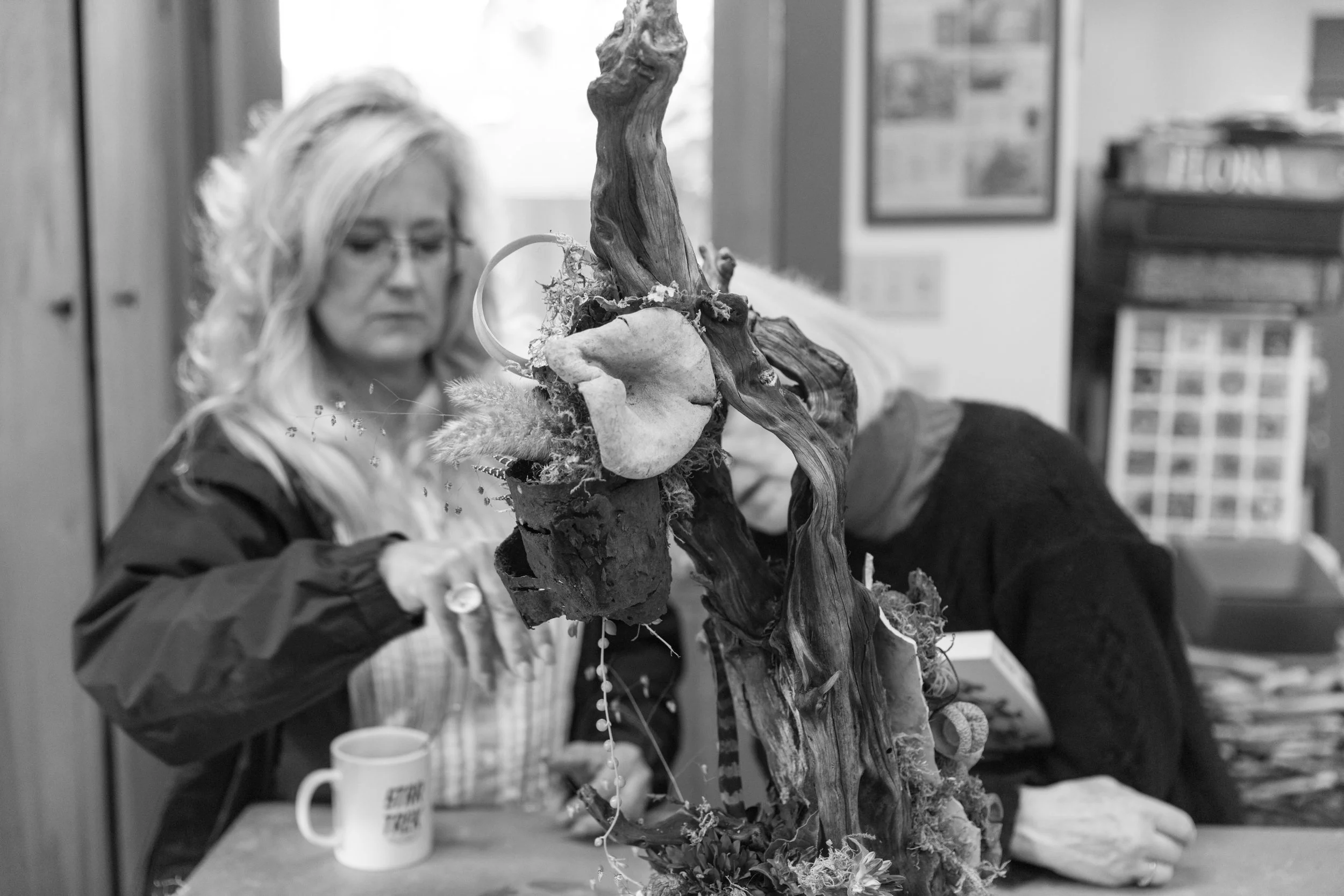 A woman with blonde curly hair, wearing glasses, is looking down in a room with a plant or floral arrangement in the foreground, and another person is partially visible next to her.