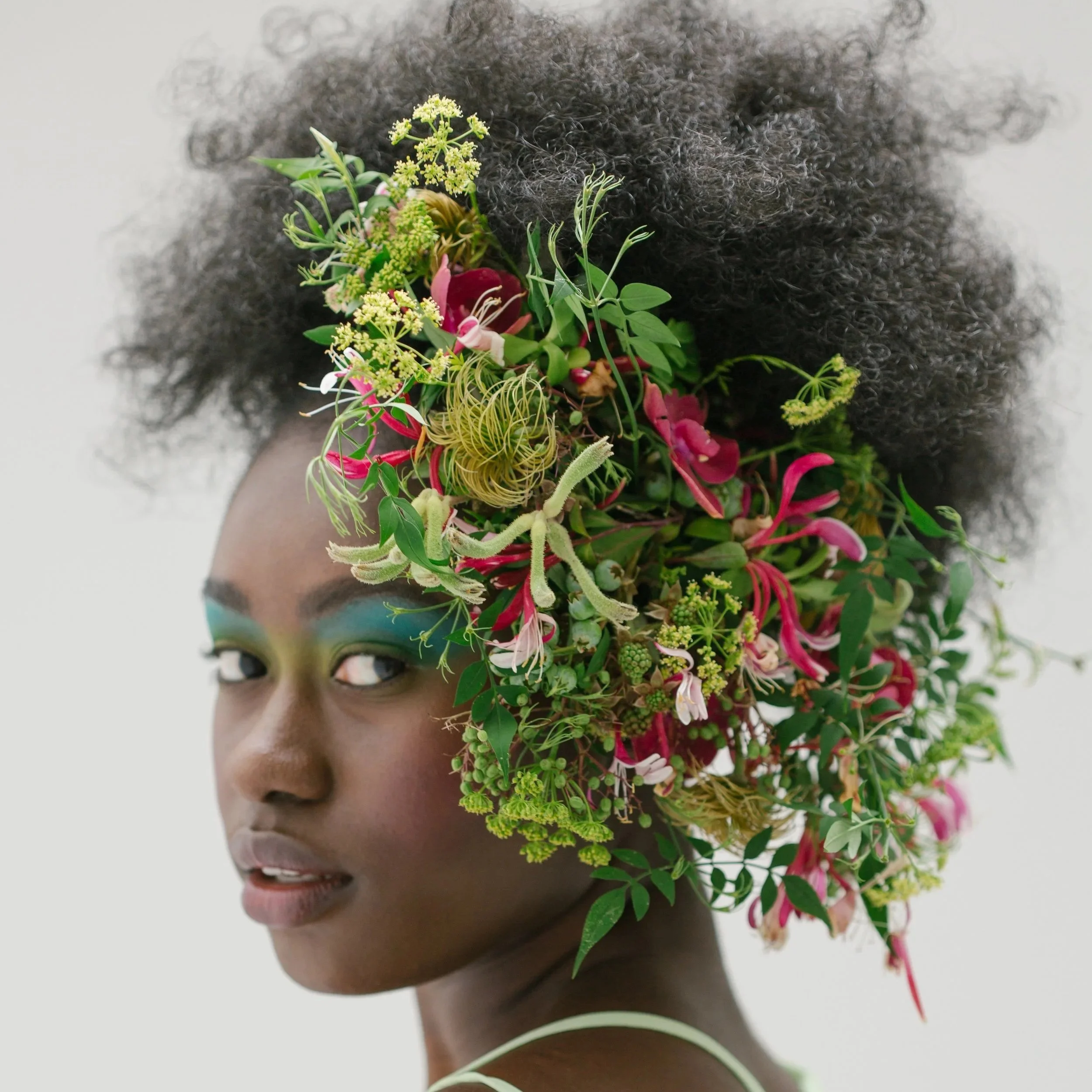 A woman with dark skin, bright makeup, and short curly hair, wearing a large floral headpiece with various green plants and pink flowers, designed by Francoise Weeks.