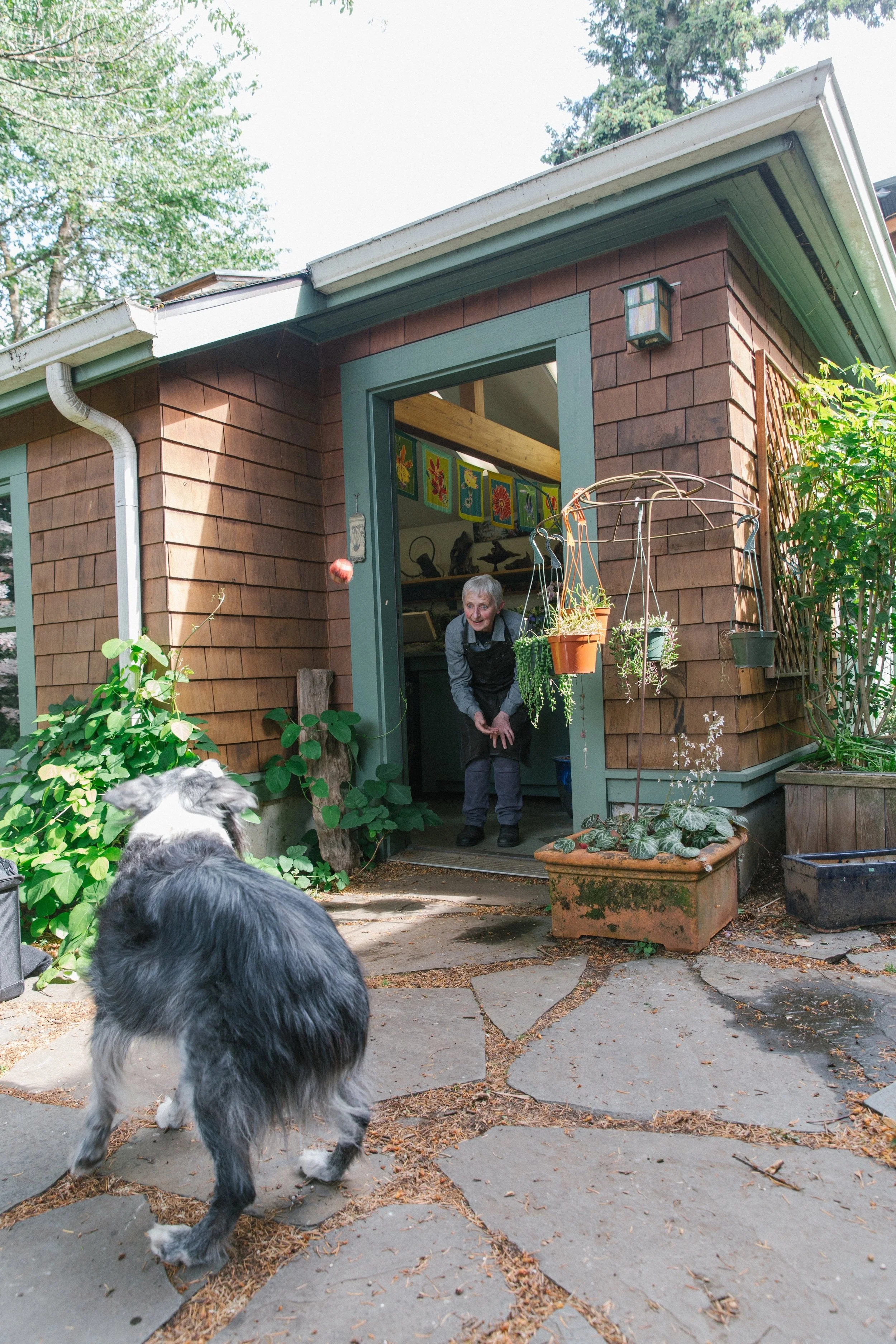 A woman is playing with a dog on a stone patio outside a wooden house, with plants and artwork visible in the background.