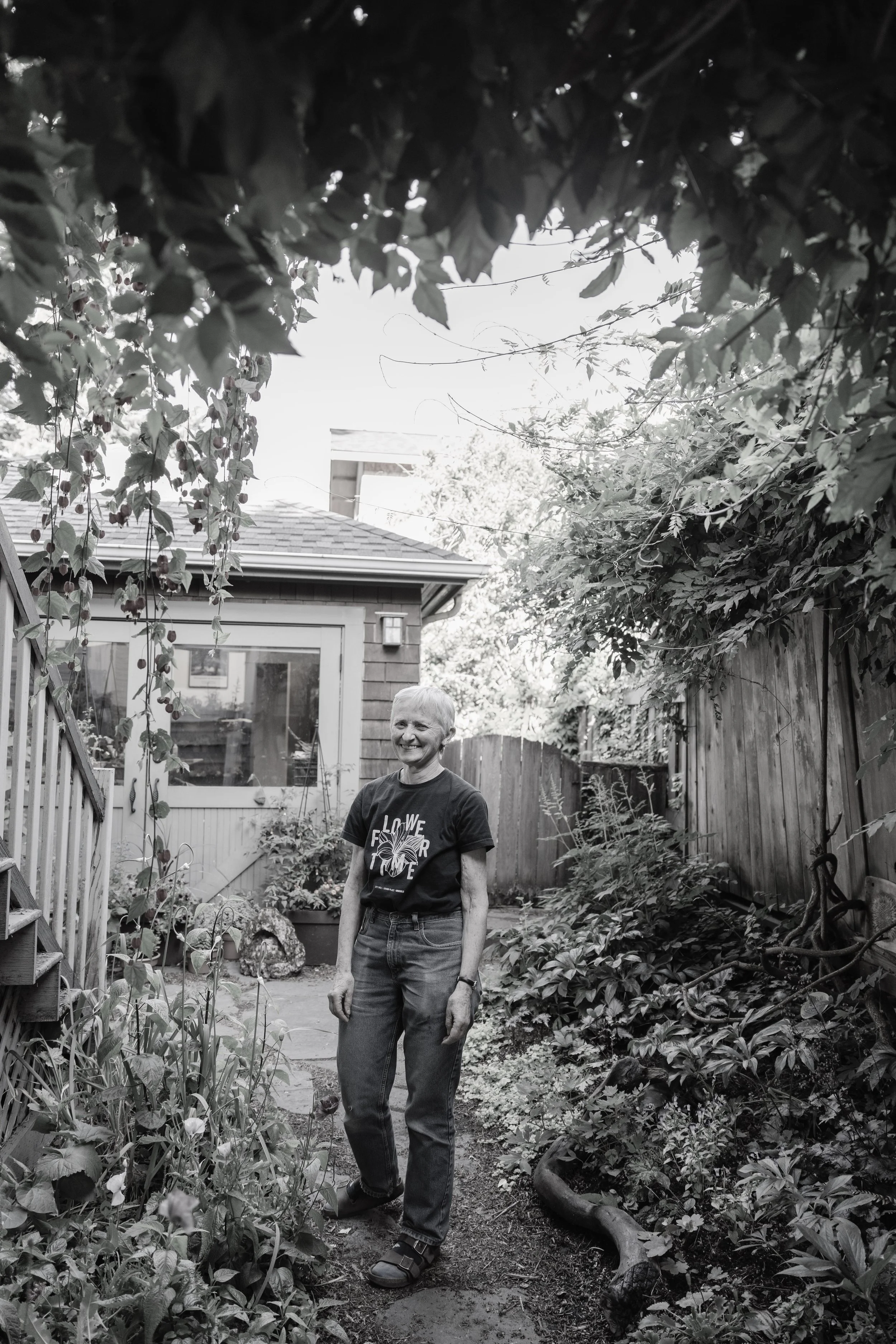 An older woman with short hair and a smile, standing in a garden yard, surrounded by plants, trees, and a wooden fence, with a house in the background.
