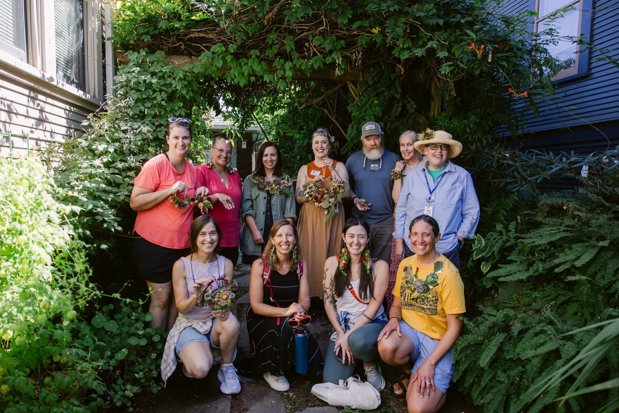 Group of ten people in a garden, some holding handmade floral arrangements, smiling for a photo under a lush green arch.