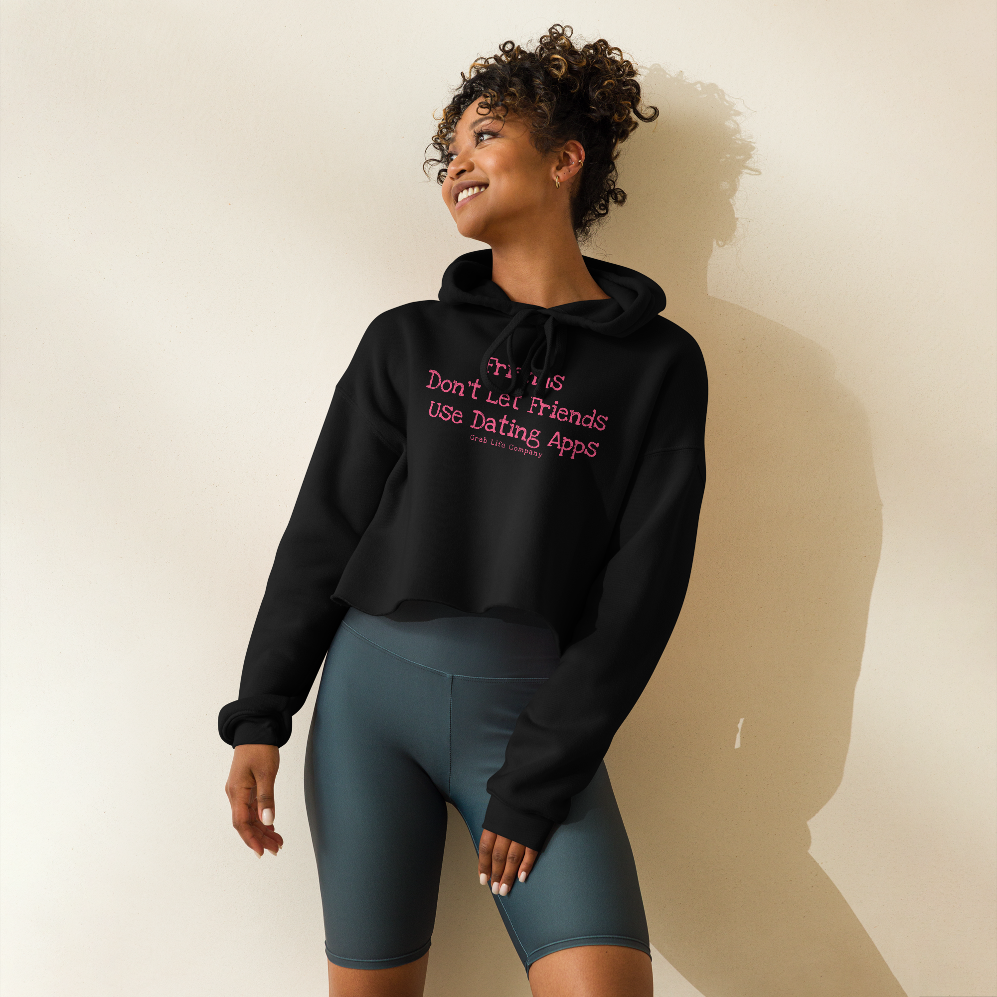 Grab Life Company cropped hoodie with Friends Don't Let Friends use Dating Apps printed on the front in pink on a black sweatshirt. Image shows a woman wearing the shirt.