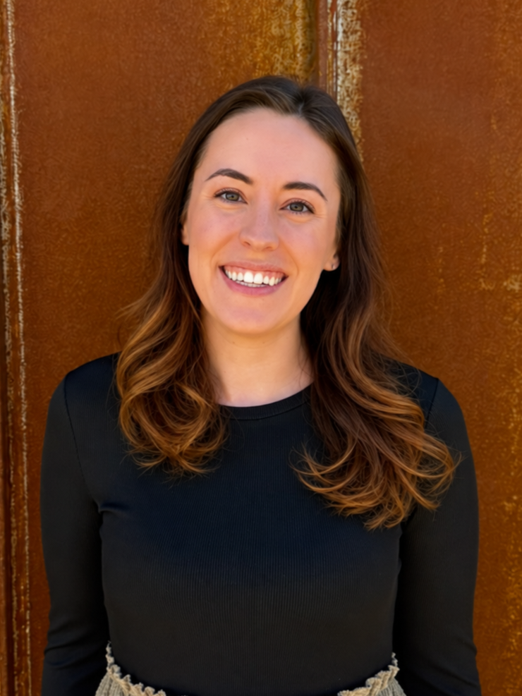 Young woman specializing in branding and web design with long wavy brown hair smiling in front of a rust-colored metal background in Colorado.