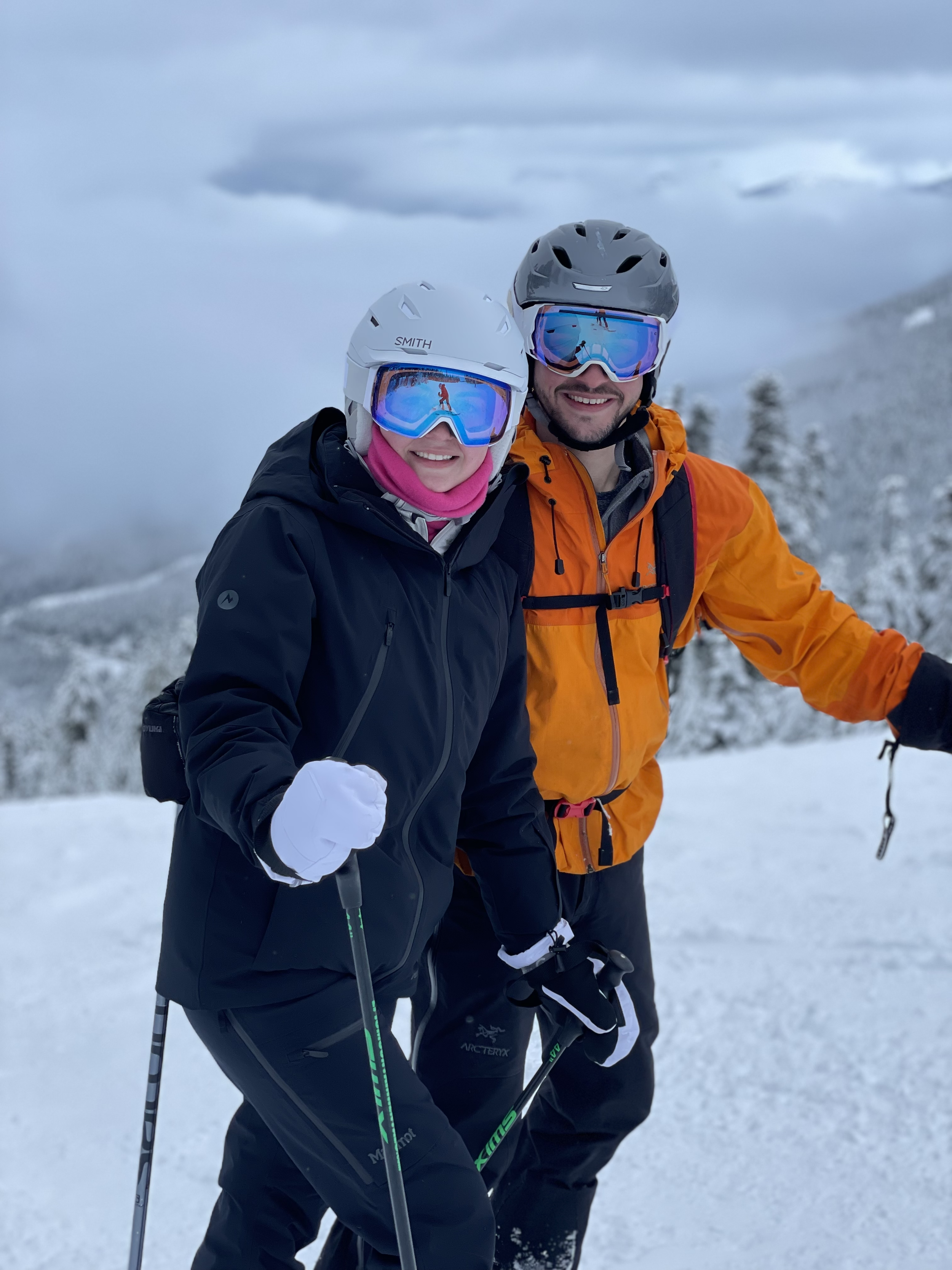 Two people in ski gear smiling on a snowy mountain with cloudy sky in the background.