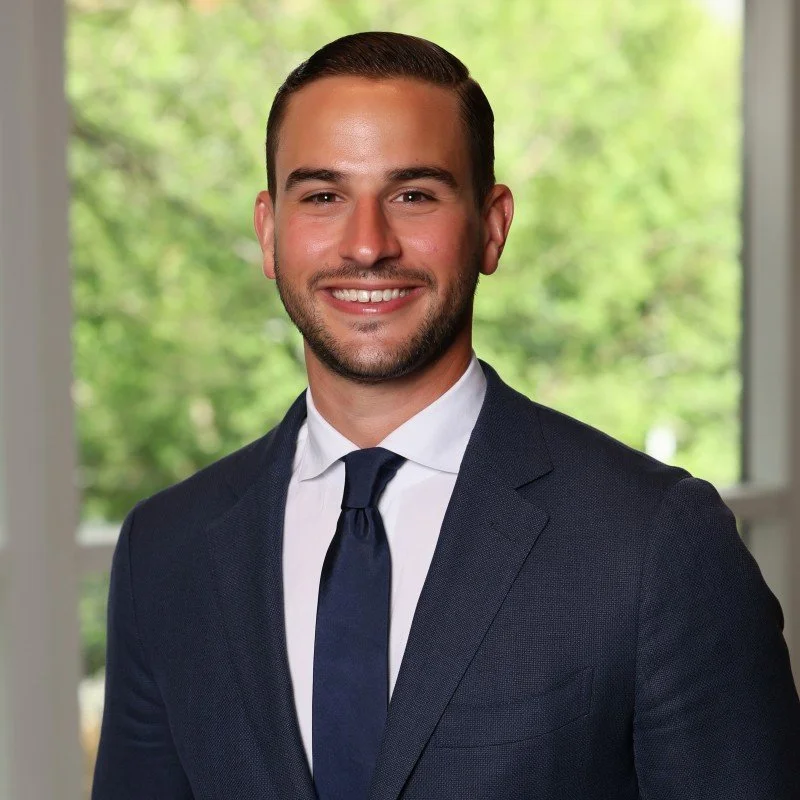 Portrait of a young man with brown hair and a beard, wearing a dark suit and tie, smiling in front of a window with greenery outside.