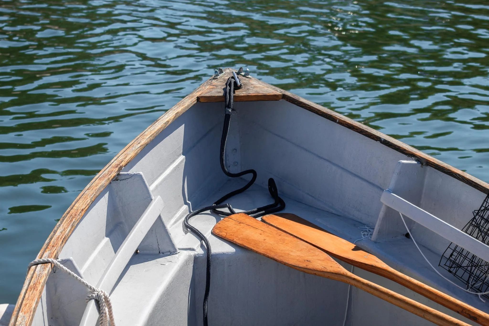 Inside of a small boat with wooden oars, black rope, and a metal wire basket, floating on water.