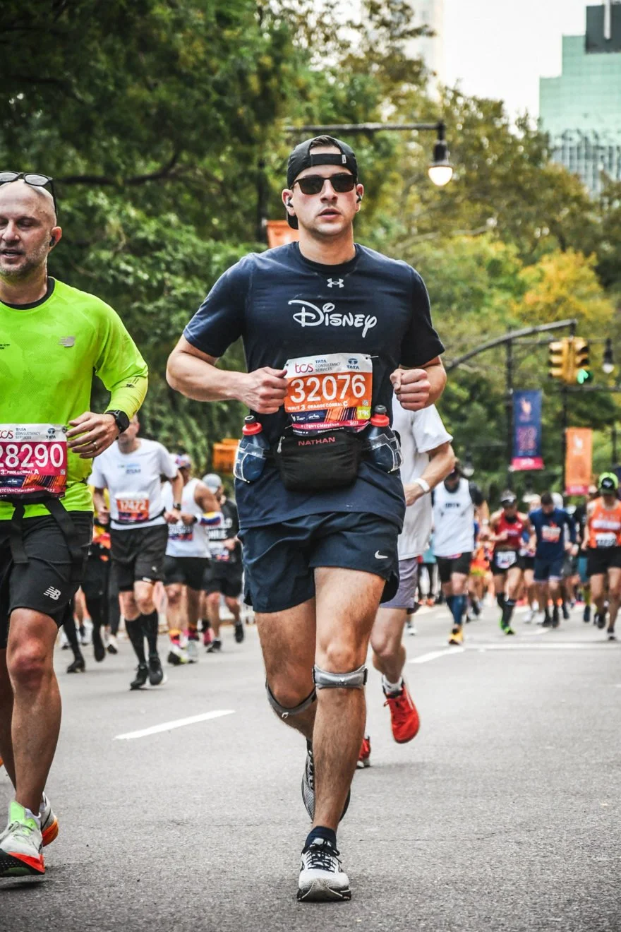 Marathon runners participating in a race in an urban setting with trees and city buildings in the background, wearing athletic gear and race bibs.