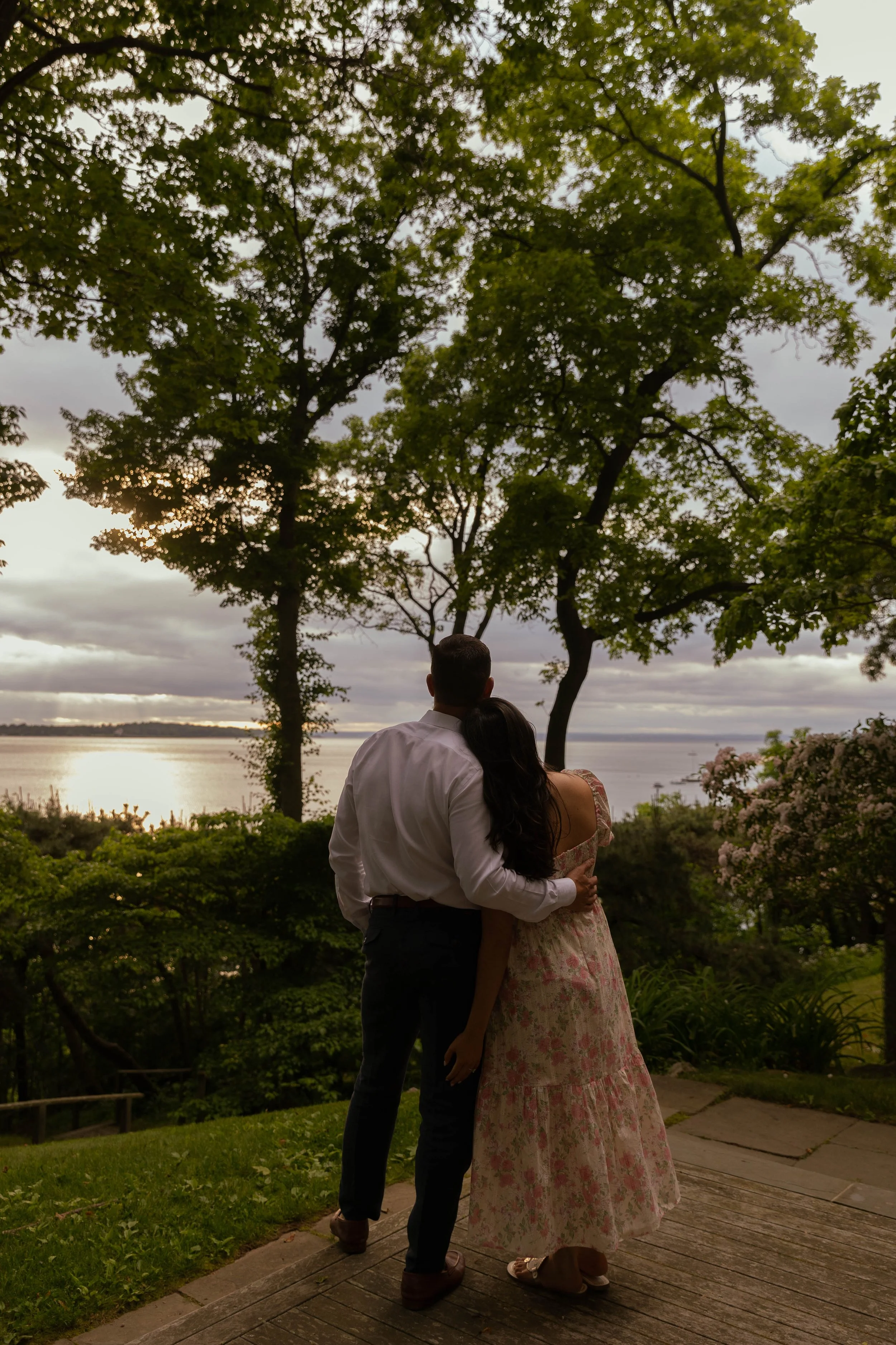 A couple standing close together on a wooden path, looking out at a lake during sunset, surrounded by trees and greenery.