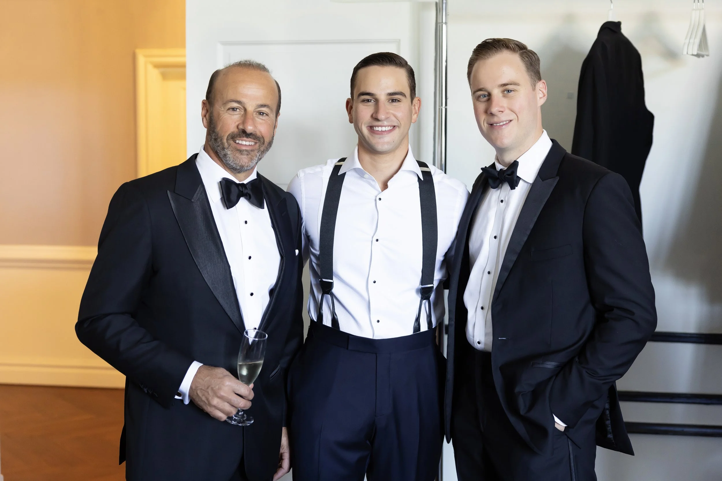 Three men in tuxedos and formal wear smiling at a gathering, one holding a glass of champagne, in a well-lit indoor space.
