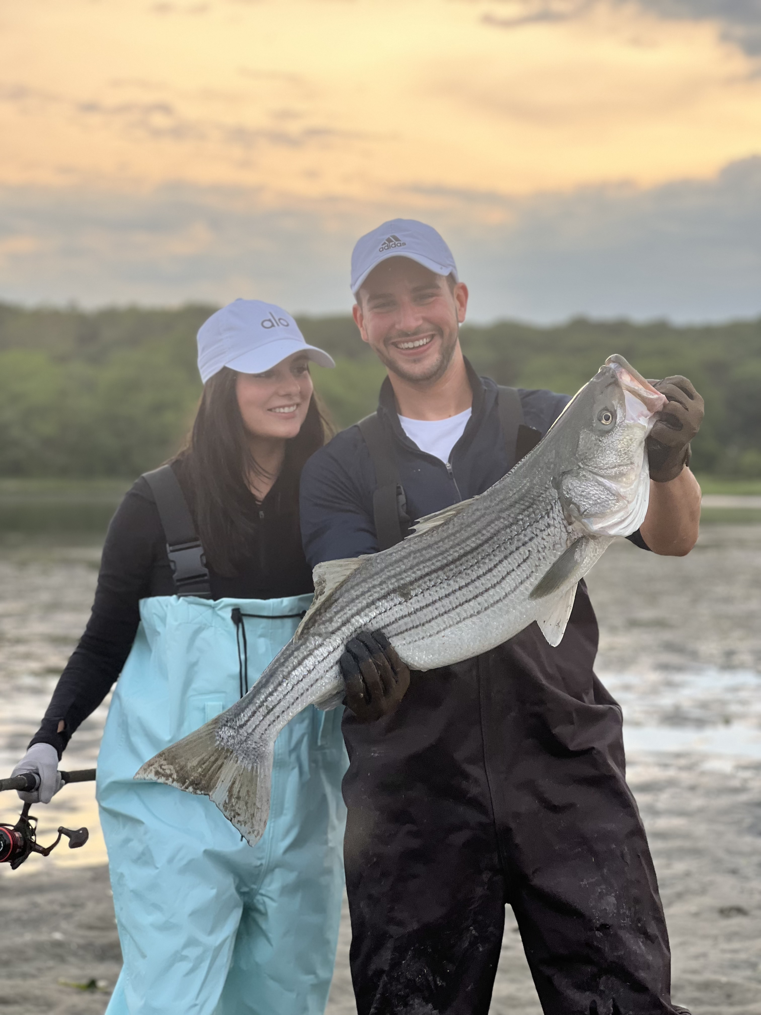 A smiling man holding a large striped bass fish outdoors at sunset with a woman nearby. Both are dressed in fishing gear and hats.