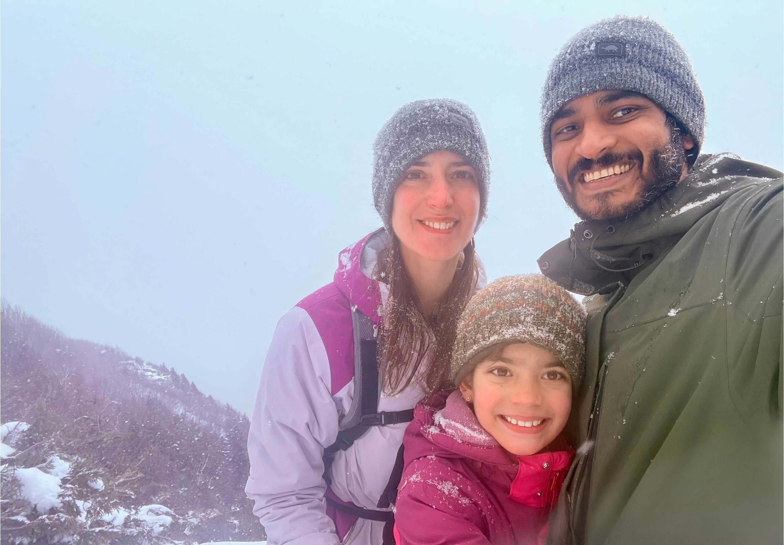 A smiling family of three taking a selfie outdoors in snowy weather, bundled in winter jackets and hats, with snow-covered trees and mountains in the background.