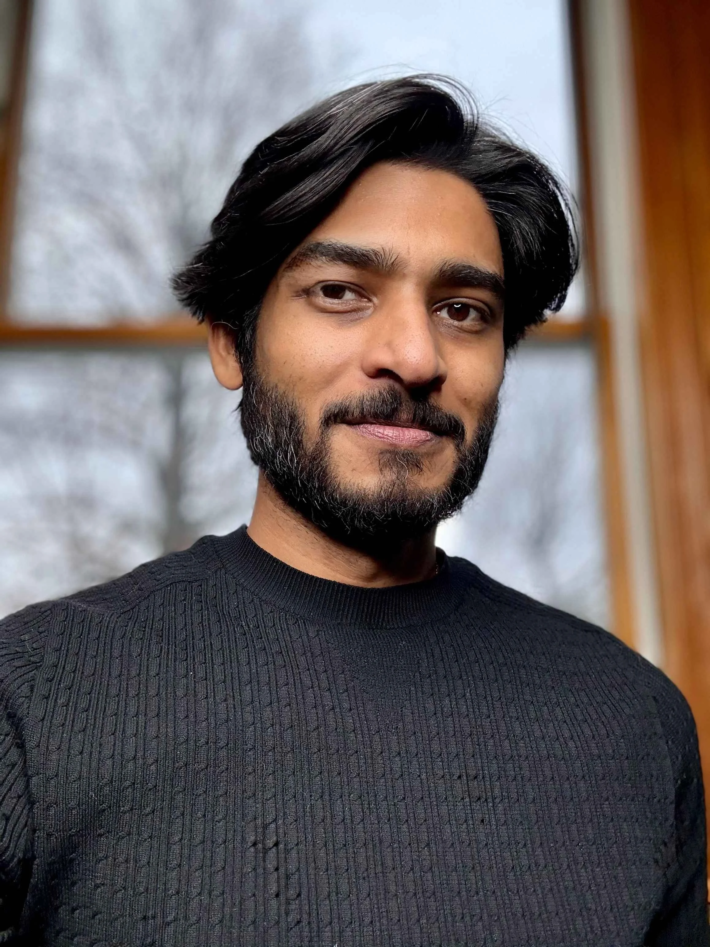 A man with black hair, a beard, and mustache, wearing a black sweater, smiling slightly, indoors with windows and trees outside in the background.