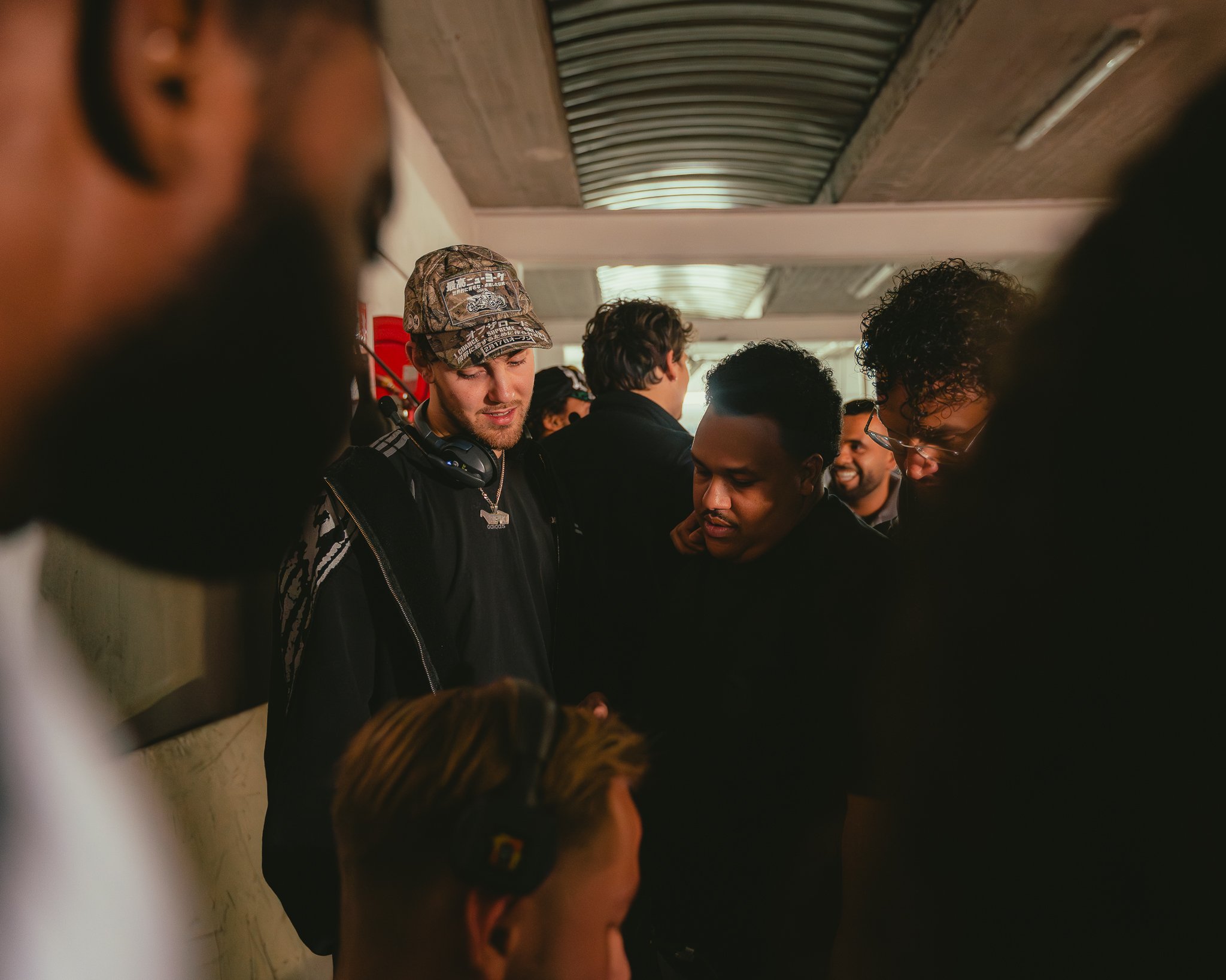 Group of men standing close together, some with headphones around their necks, in an indoor setting with a corrugated metal ceiling.