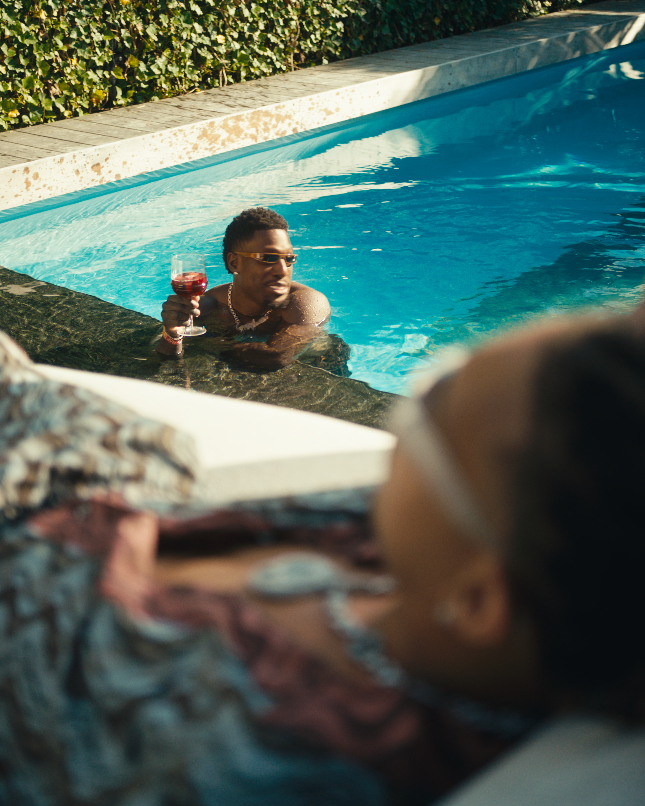 A man with sunglasses and jewelry relaxing in a swimming pool, holding a glass of red wine, with lush green foliage and a stone border in the background.