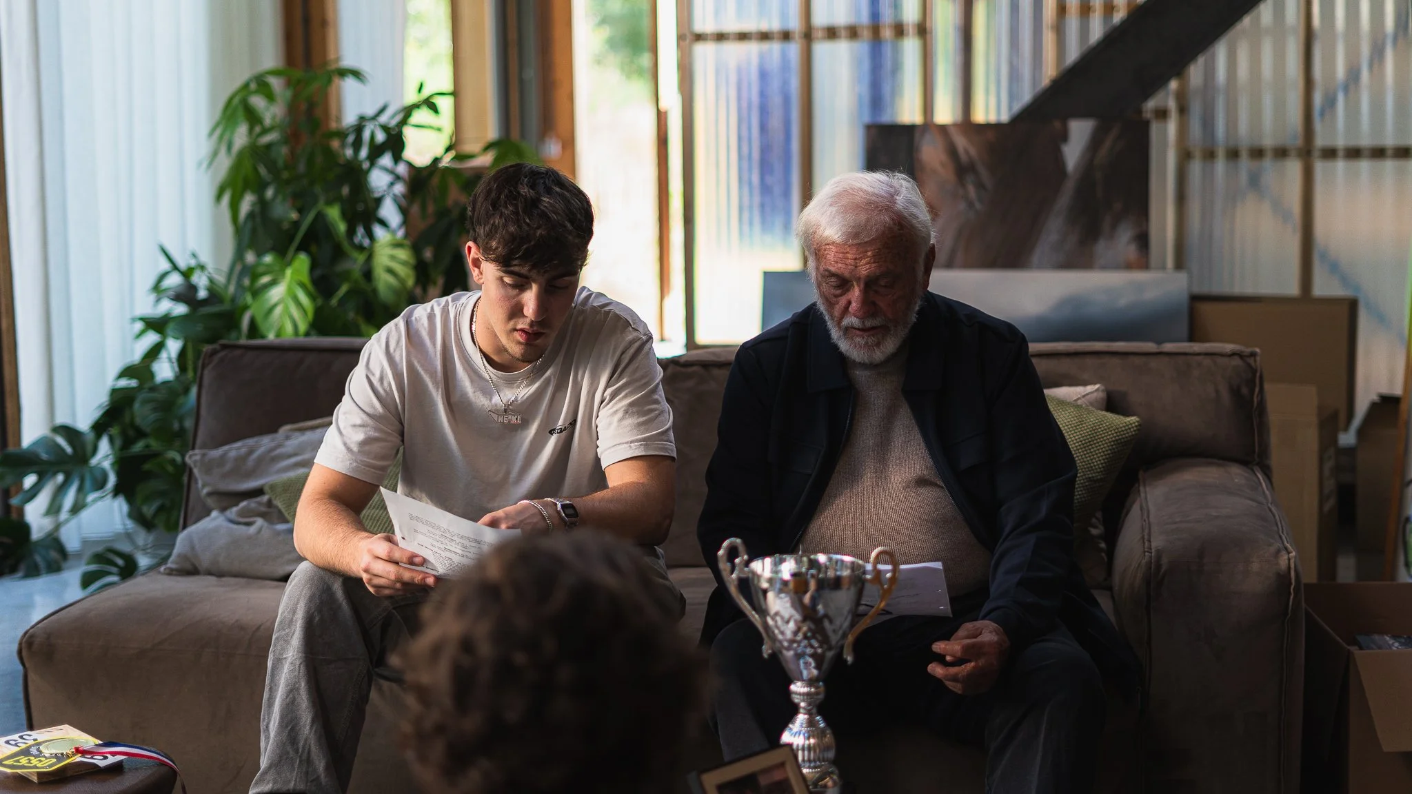 A young man and an older man sitting on a sofa, reading papers, with a trophy on the table in front of them, in a cozy room with large windows, plants, and a staircase in the background.