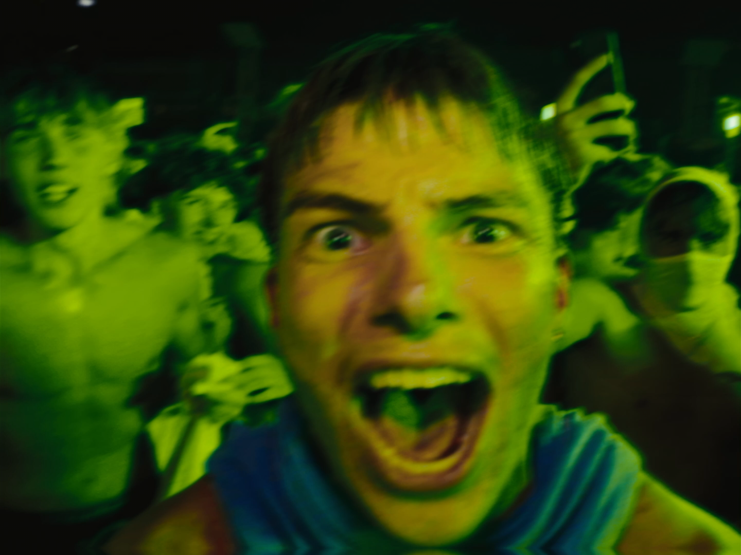 A group of people, with a man in the foreground shouting or yelling, with an intense expression, surrounded by others, some wearing masks, all illuminated with green lighting. yeat concert