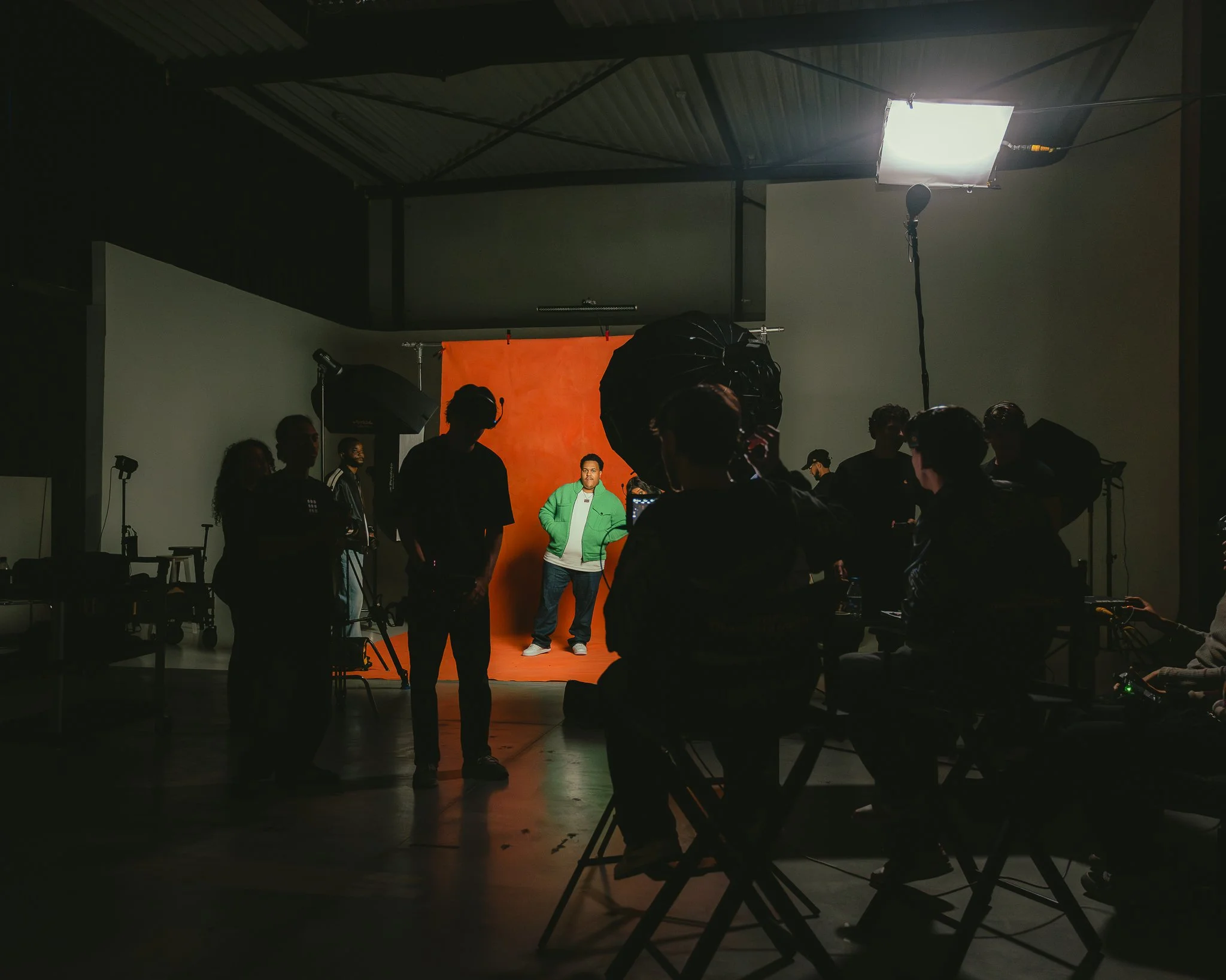 People working on a photoshoot with a man in a green jacket posing in front of an orange backdrop, inside a studio with professional lighting and equipment.
