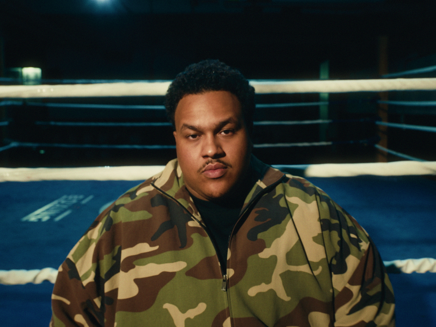 A young man in a camouflage jacket standing in a boxing ring at night.