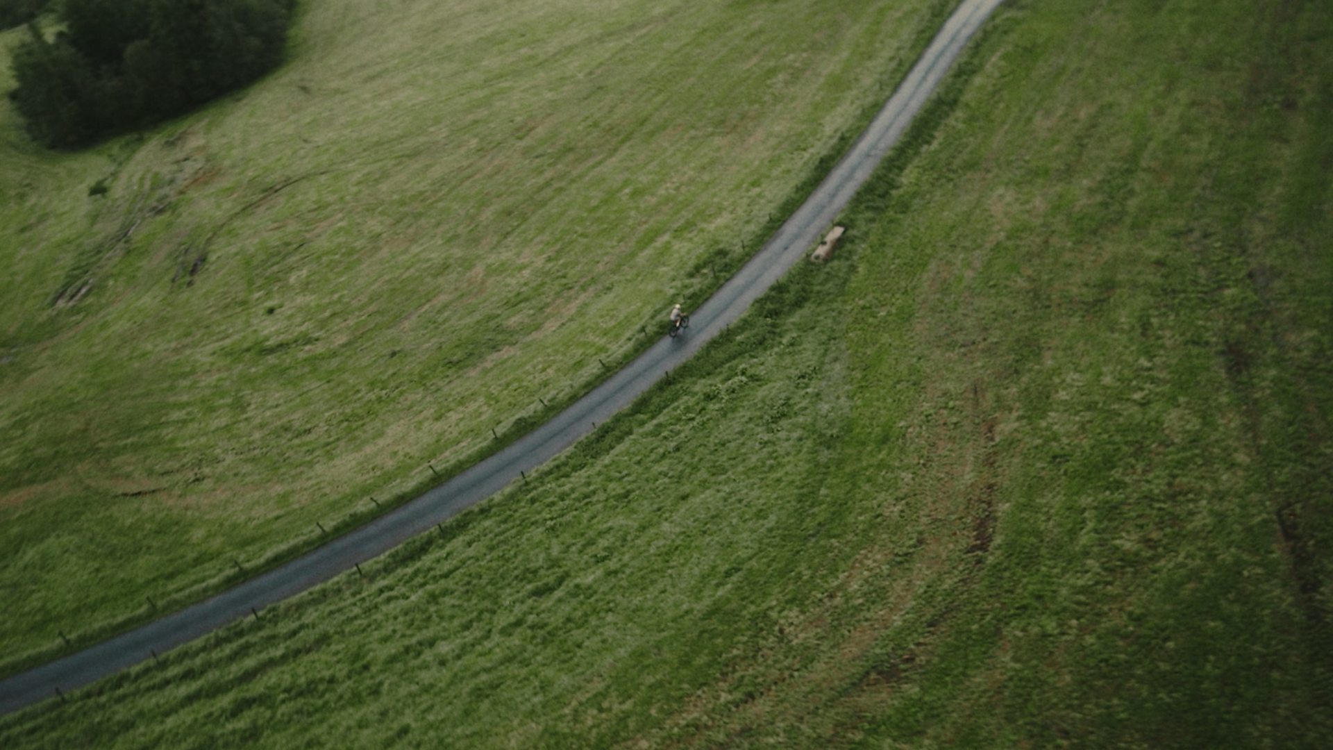 A person riding a bicycle on a narrow, winding paved path through green grassy fields, with a few scattered trees in the background.
