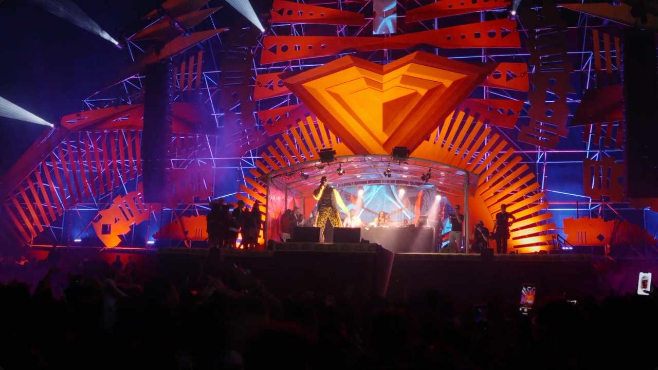 Stage with bright orange and purple lighting, large geometric logo at the top, performer in colorful outfit at the center, and live band with audience in foreground.