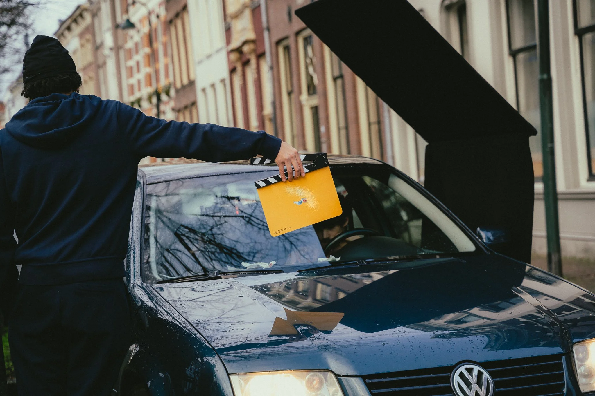 A person holding a movie clapperboard next to a black Volkswagen car with its hood open, parked on a city street with colorful buildings in the background. Heki.made heki collective film