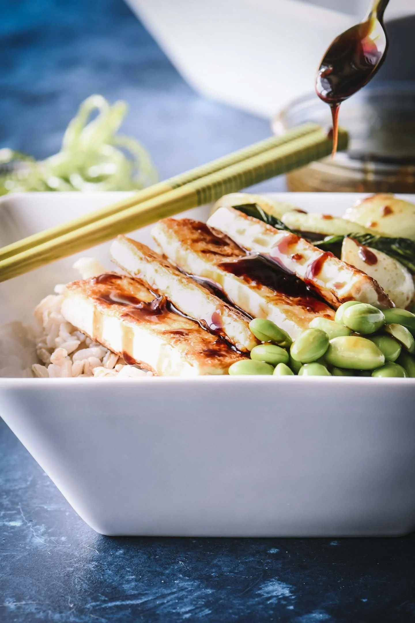 A bowl of sliced tofu topped with a dark sauce, accompanied by edamame, chopped vegetables, and green onions, with a spoon drizzling sauce over the tofu.