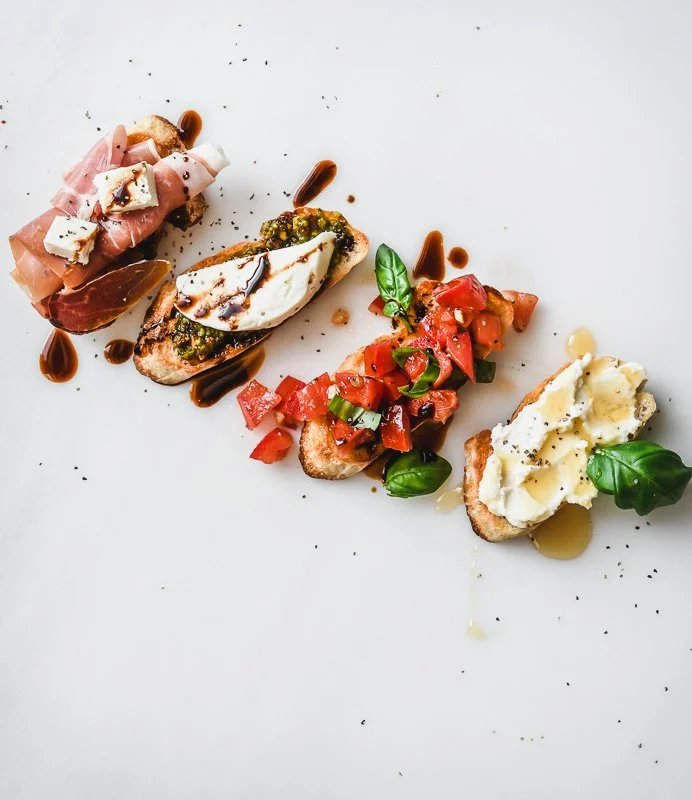 Four bruschetta toasts decorated with various toppings, including prosciutto and cheese, basil and tomato, and potato salad, on a white surface.