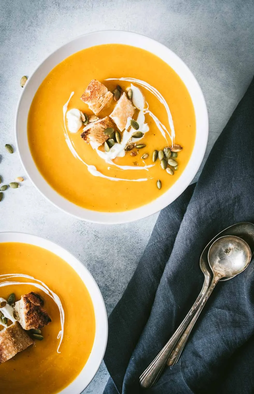 Two bowls of creamy, orange soup garnished with croutons, pumpkin seeds, and a swirl of cream on top, placed on a gray surface with silver spoons and a dark napkin.