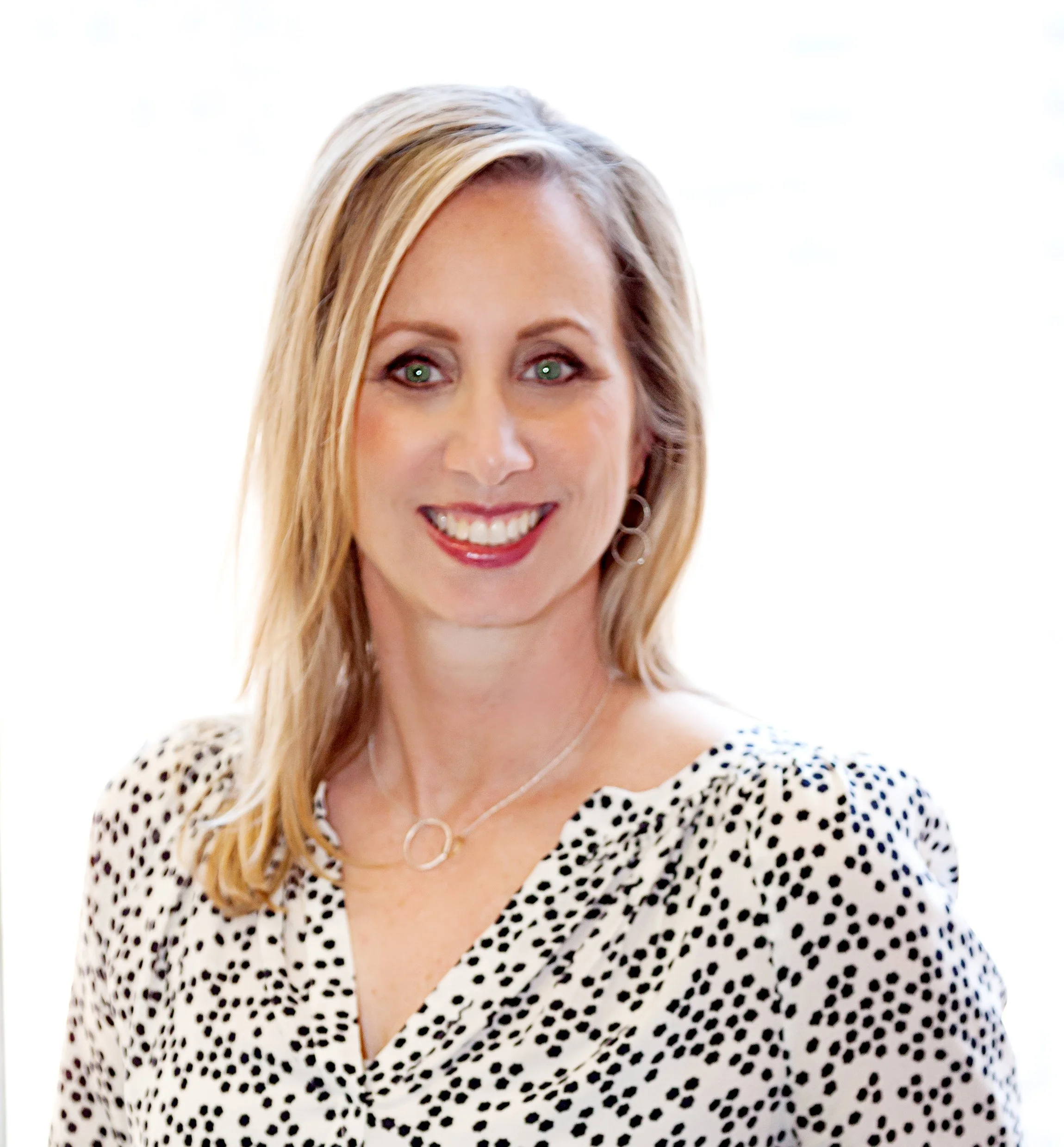 A smiling woman with blonde hair, green eyes, wearing a black and white polka dot top, silver jewelry, and earrings, against a bright background.